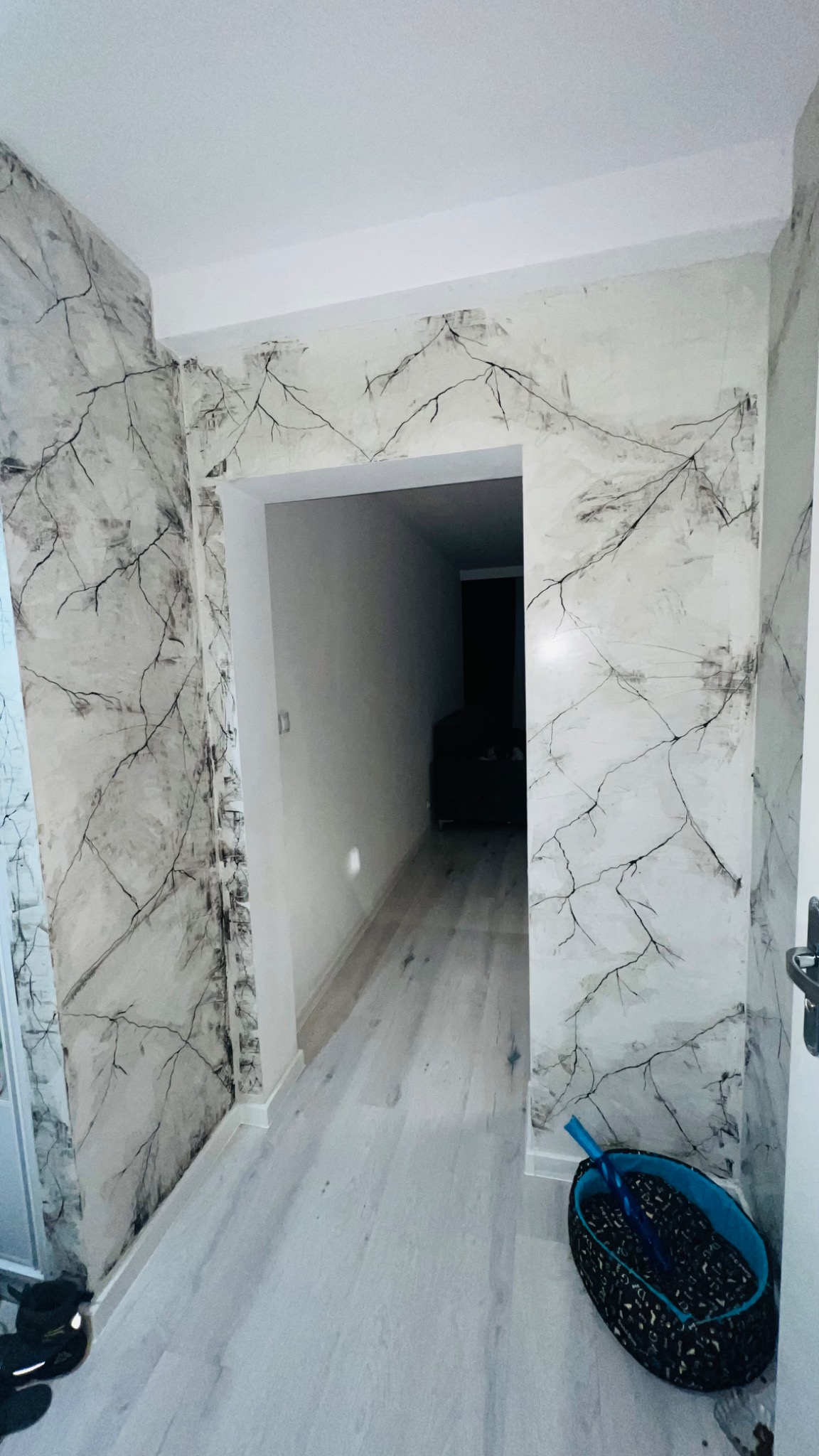 Korytarz po remoncie z dekoracyjną, szaro-białą ścianą imitującą marmur z czarnymi żyłkami, widoczne wejście do pokoju i kosz z narzędziami malarskimi.