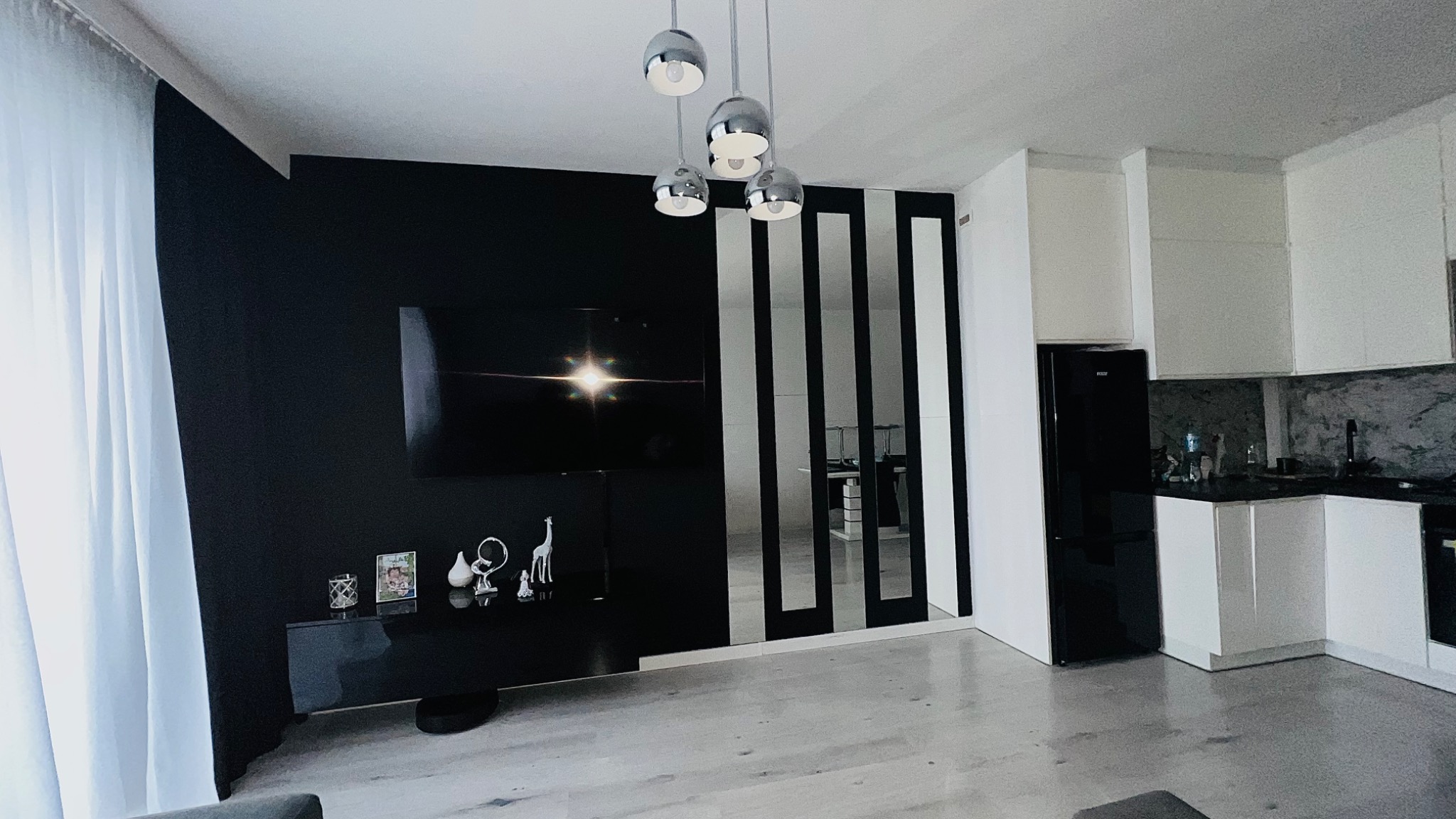 Nowoczesne wnętrze salonu z czarną ścianą, telewizorem, dekoracjami, lustrami w czarnych ramach i widokiem na kuchnię z białymi szafkami i czarną lodówką.