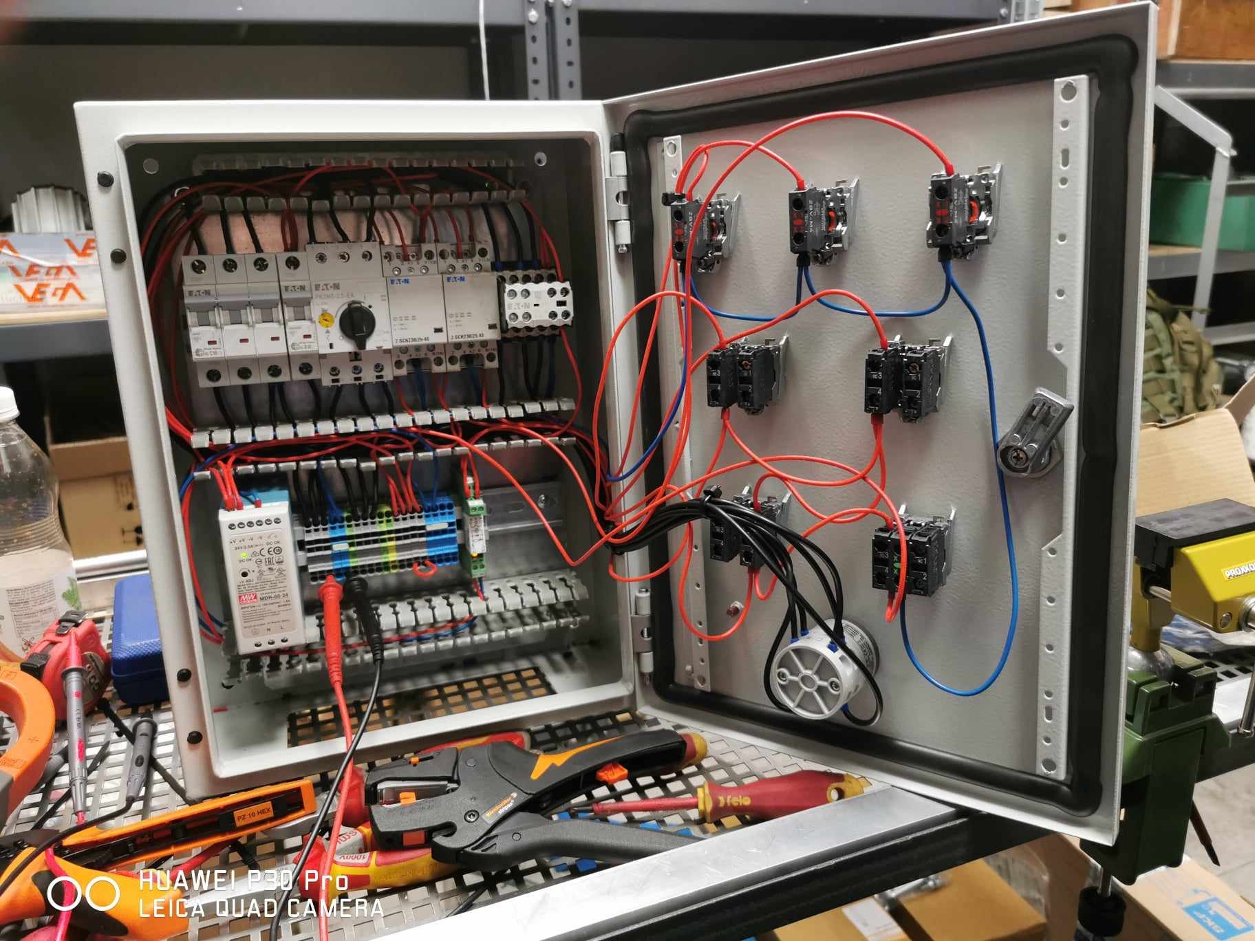 Otwarta skrzynka elektryczna z widocznymi bezpiecznikami, przewodami w kolorach czerwonym i niebieskim oraz narzędziami elektrycznymi na dole, w tym miernik i kombinerki.