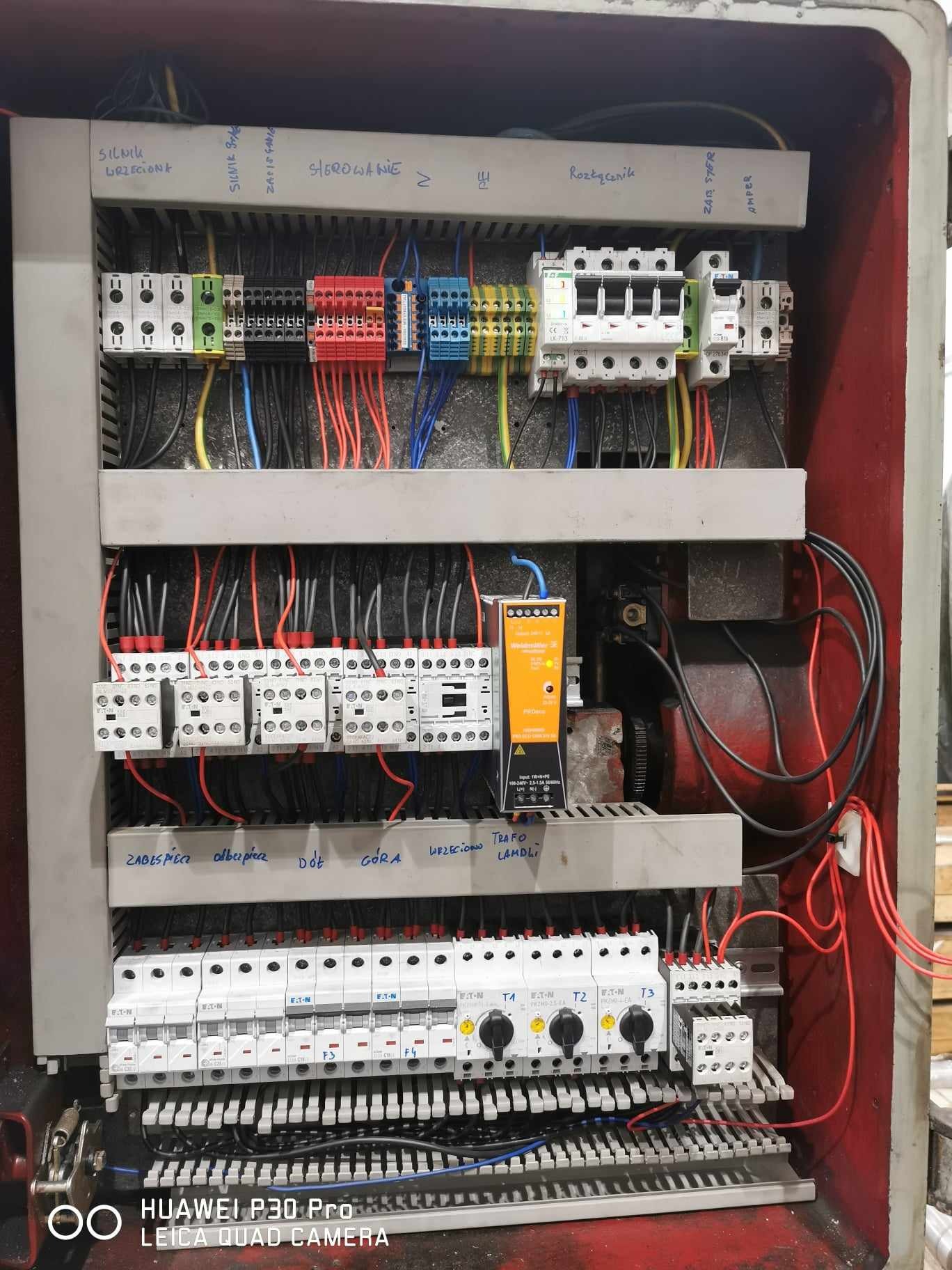Otwarta szafa sterownicza z widocznymi elementami instalacji elektrycznej, w tym bezpieczniki, przekaźniki i okablowanie, opisane ręcznie na tabliczkach, wewnątrz obudowy pomalowanej na czerwono.