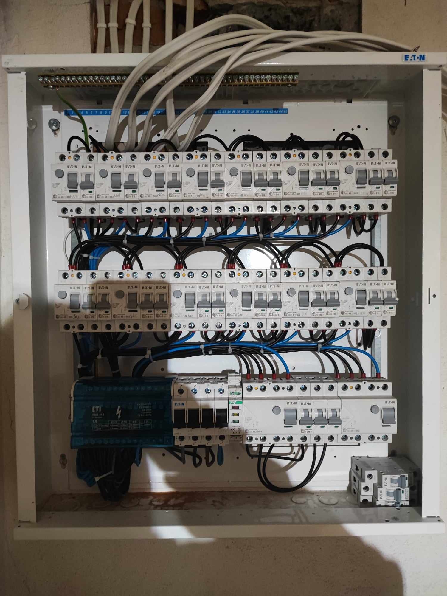 Rozdzielnica elektryczna z zainstalowanymi bezpiecznikami EATON i przewodami w kolorach czarnym i niebieskim, widoczne elementy montażowe i oznaczenia na obudowie.
