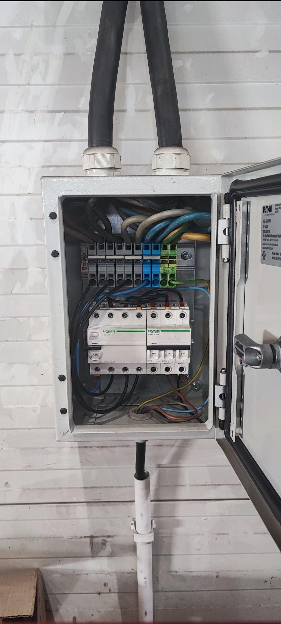 Otwarta skrzynka elektryczna z bezpiecznikami Schneider Electric i przewodami w różnych kolorach, zamontowana na białej ścianie z boazerii.