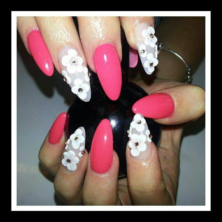 Ręka z pomalowanymi paznokciami: trzy różowe, gładkie paznokcie i dwa z białymi kwiatami 3D z drobnymi złotymi koralikami pośrodku, na tle czarnego, błyszczącego przedmiotu.