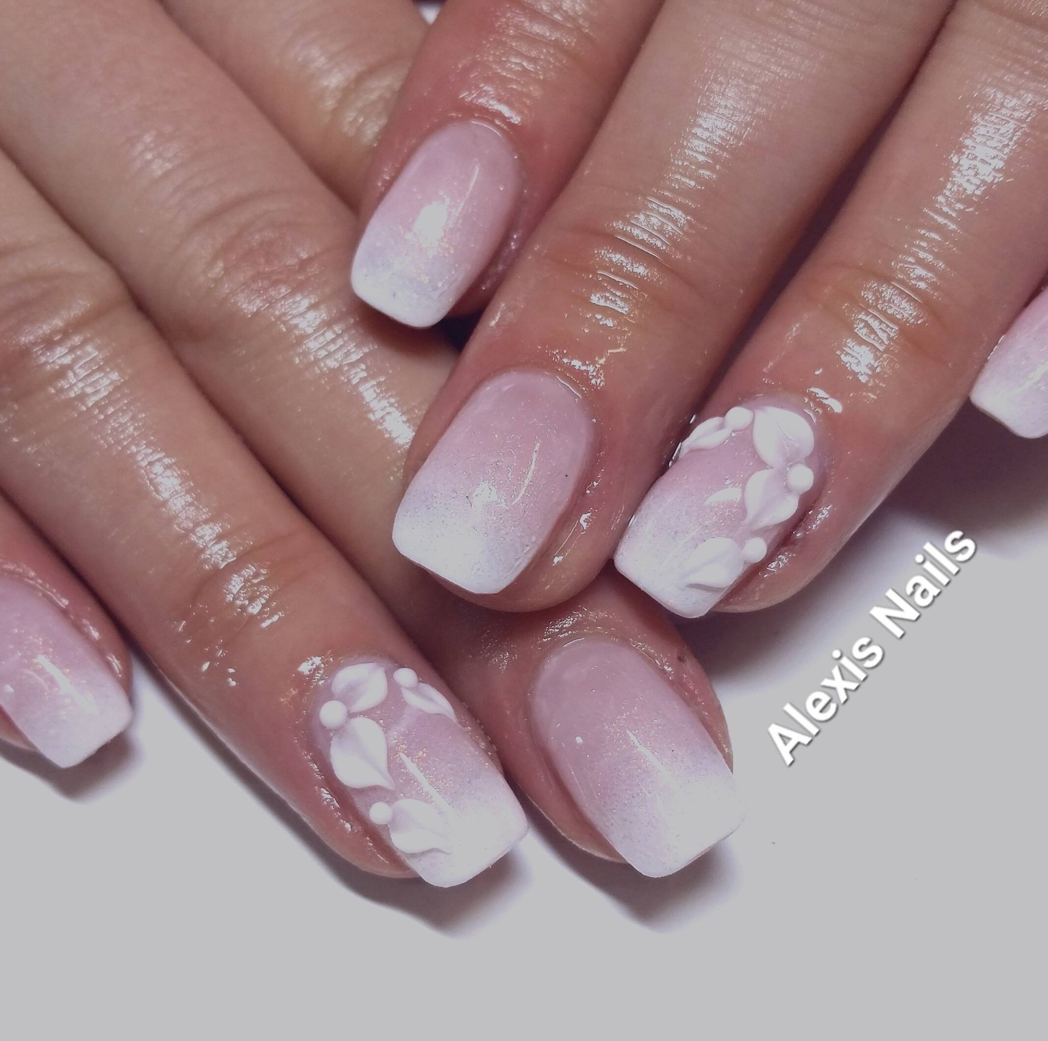 Dłonie z manicure hybrydowym ombre w odcieniach różu i bieli, zdobione białymi, wypukłymi wzorami kwiatowymi na dwóch paznokciach.