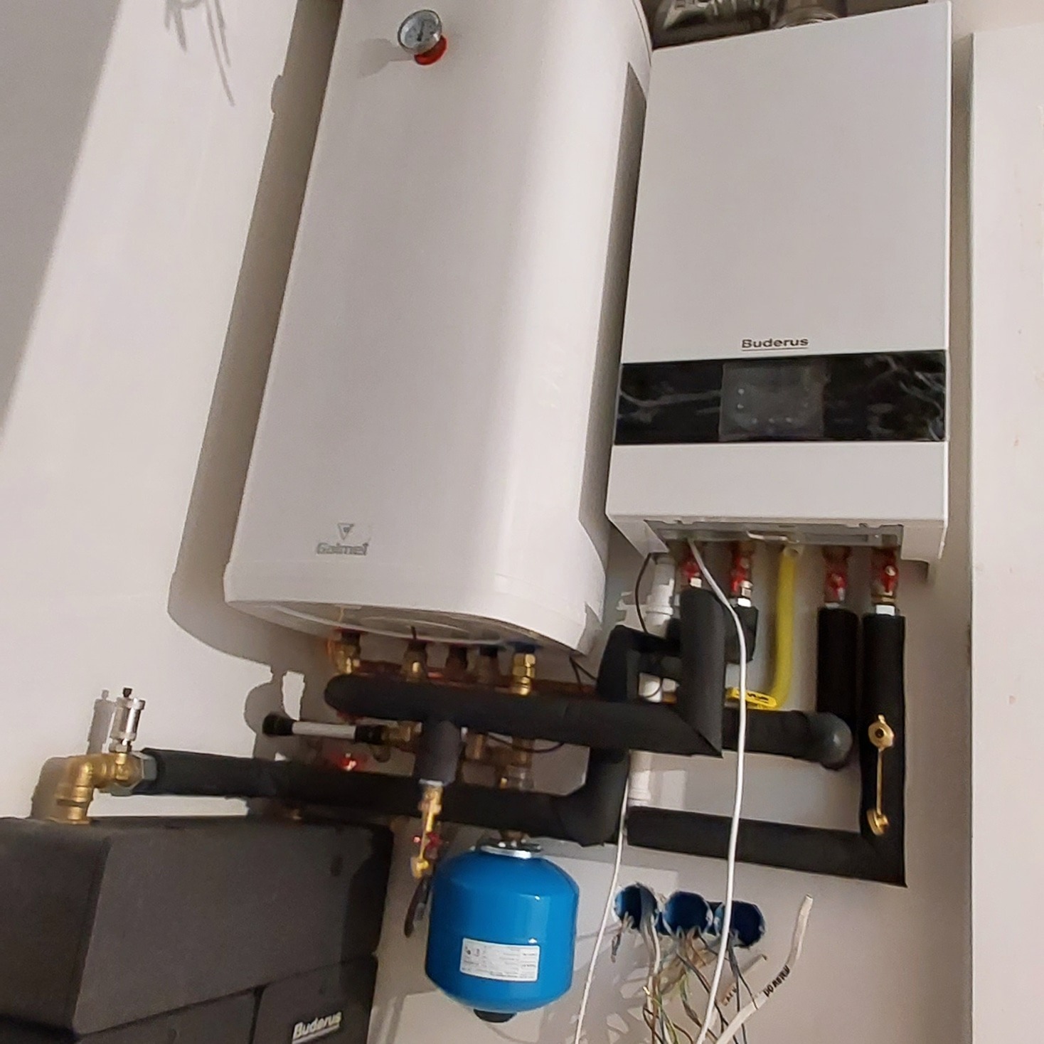 Biały zasobnik ciepłej wody użytkowej Galmet i wiszący kocioł gazowy Buderus z podłączeniami rur w izolacji, niebieski zbiornik przeponowy i widoczne przewody elektryczne.