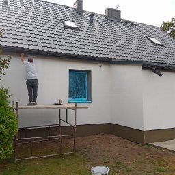 F.U. Mirax Mirosław Grzesiak - Pracownik na rusztowaniu wyrównuje świeżo otynkowaną elewację domu jednorodzinnego, okna zabezpieczone folią, widoczny fragment dachu z dachówką w kolorze grafitowym.
