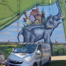 F.U. Mirax Mirosław Grzesiak - Duży mural na ścianie budynku przedstawiający słonia niosącego na grzbiecie miasto; na pierwszym planie srebrny samochód dostawczy i drabina oparta o ścianę, obok dwa wózki narzędziowe.