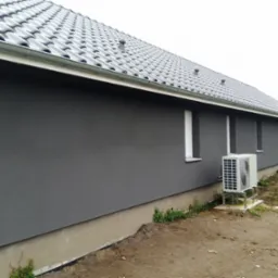 Szara elewacja budynku jednorodzinnego z widoczną jednostką zewnętrzną klimatyzacji, szarym dachem ceramicznym i metalową rynną, trawnik w trakcie zakładania.