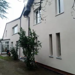 Jasna elewacja domu jednorodzinnego po tynkowaniu, widoczne okna z szarymi parapetami, czarne rynny i fragment ogrodu z zielenią