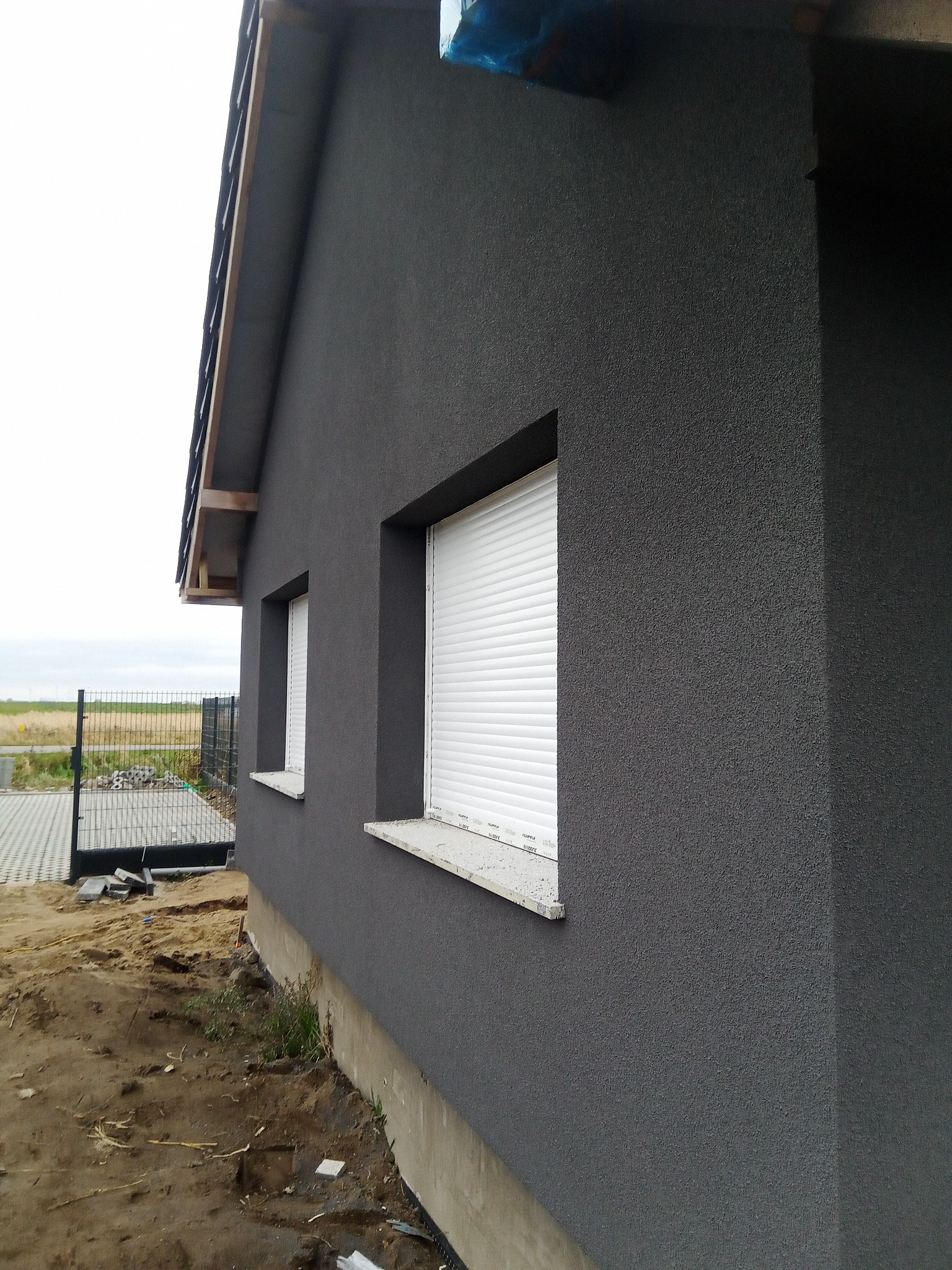 Świeżo otynkowana, grafitowa elewacja domu jednorodzinnego z białymi roletami zewnętrznymi, widoczny fragment dachu i teren wokół budynku w trakcie prac wykończeniowych.