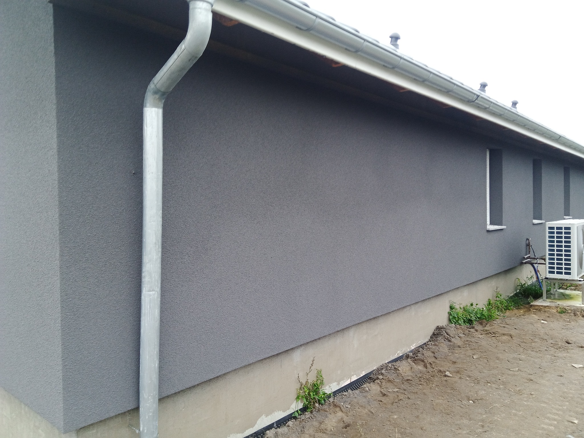 Szara elewacja budynku jednorodzinnego z widoczną rynną, fragmentem dachu i klimatyzatorem na zewnątrz