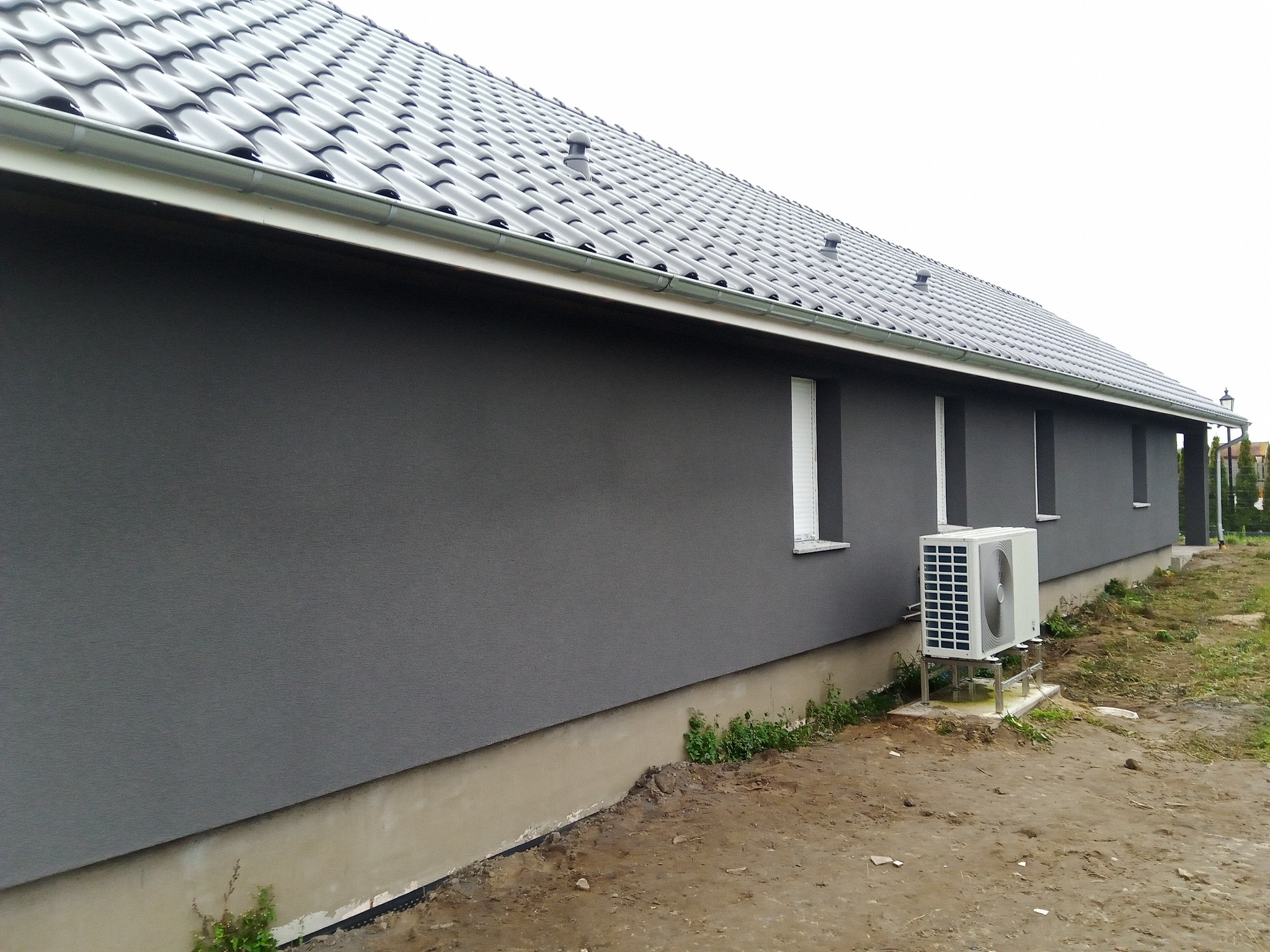 Szara elewacja budynku jednorodzinnego z widoczną jednostką zewnętrzną klimatyzacji, szarym dachem ceramicznym i metalową rynną, trawnik w trakcie zakładania.