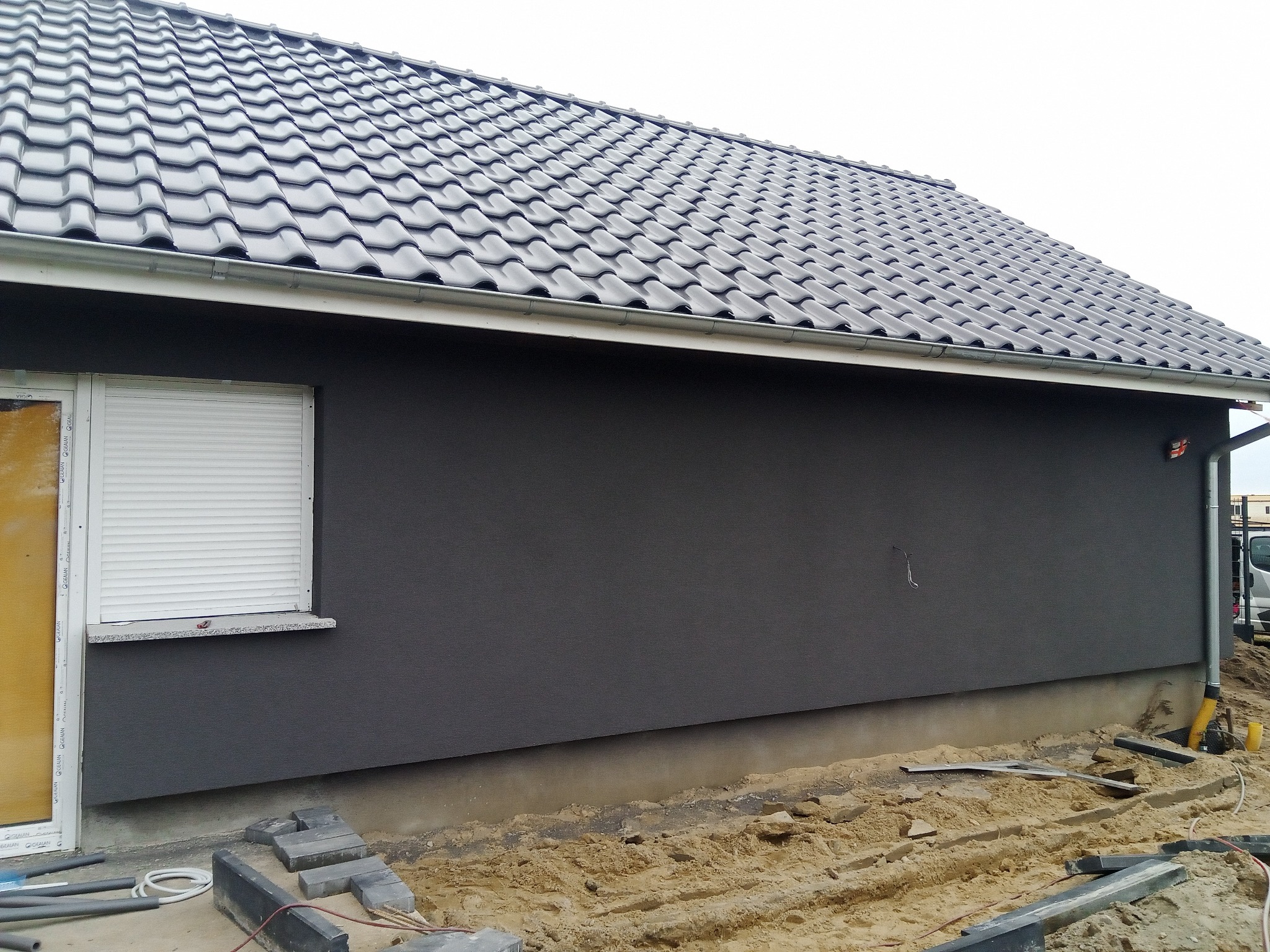 Ściana domu z ciemnoszarą elewacją i oknem z żaluzjami zewnętrznymi, widoczny fragment dachu z szarą dachówką i teren wokół domu w trakcie prac wykończeniowych.