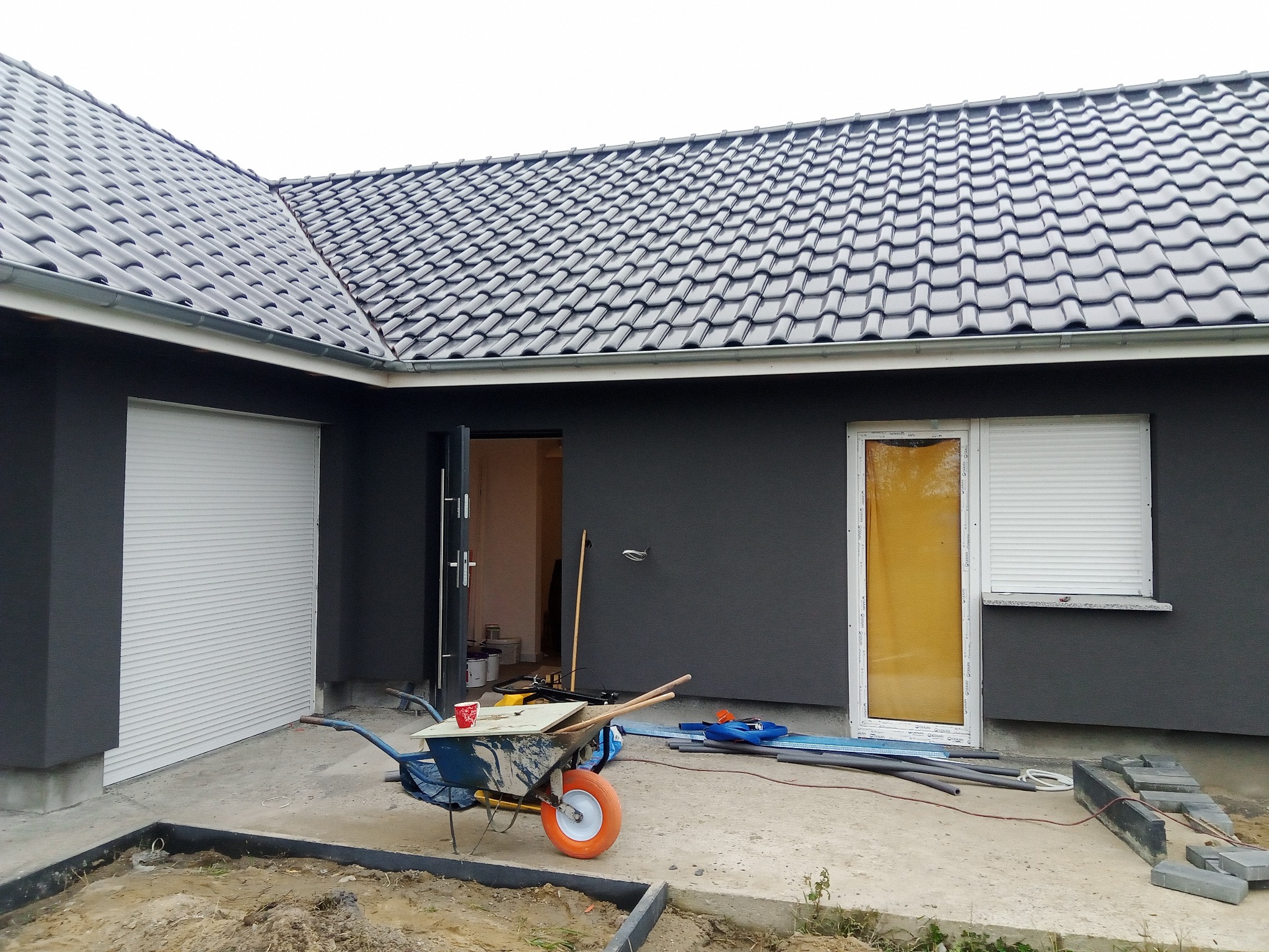 Dom z szarą elewacją i ciemnoszarą dachówką w trakcie prac wykończeniowych, widoczny brudny wózek budowlany z narzędziami i kubkiem, okno z roletą i fragmenty kostki brukowej.