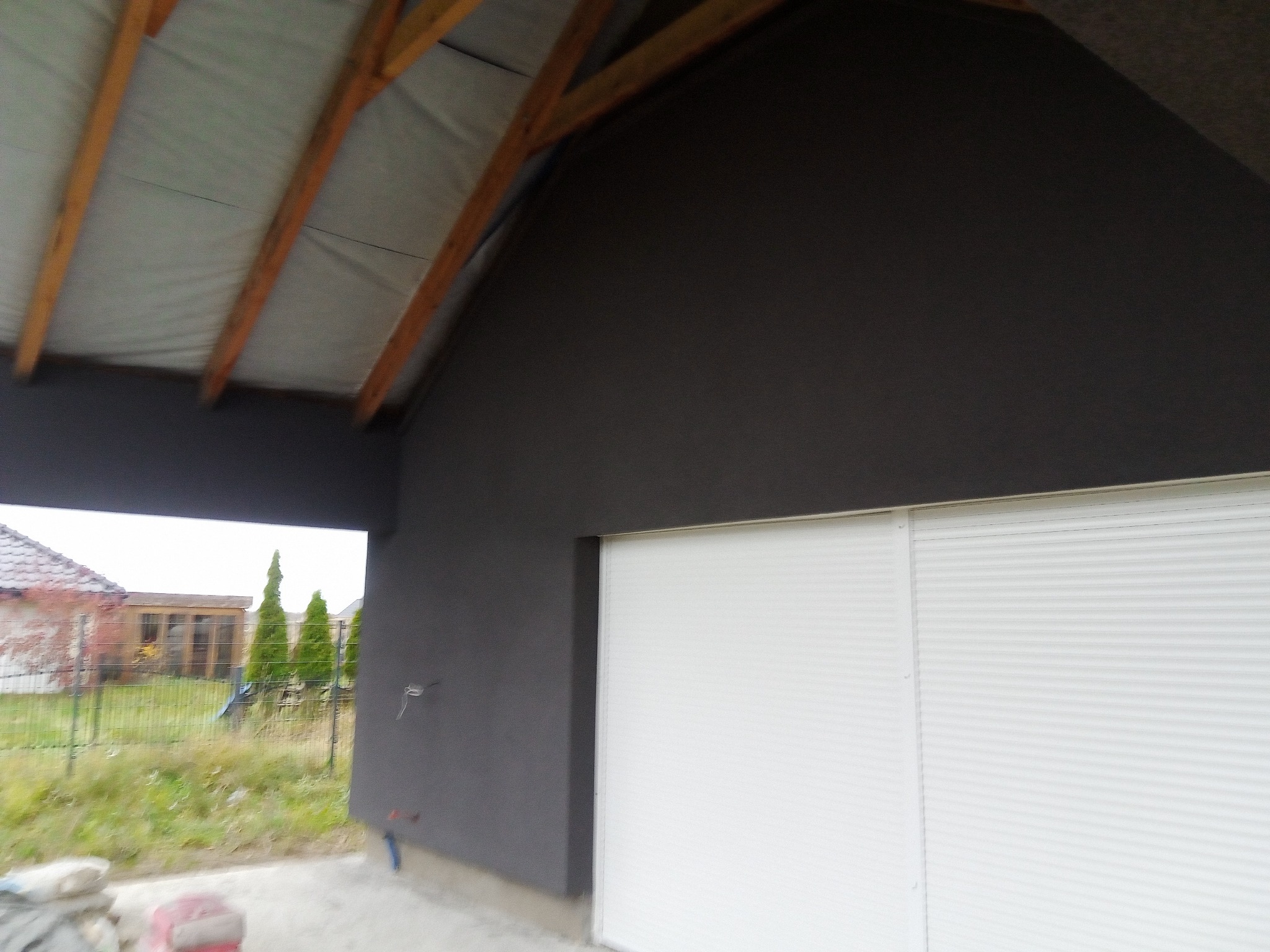 Świeżo otynkowana, ciemnoszara elewacja budynku z białymi roletami zewnętrznymi, widok z zewnątrz, w tle zieleń ogrodu i sąsiedni budynek.