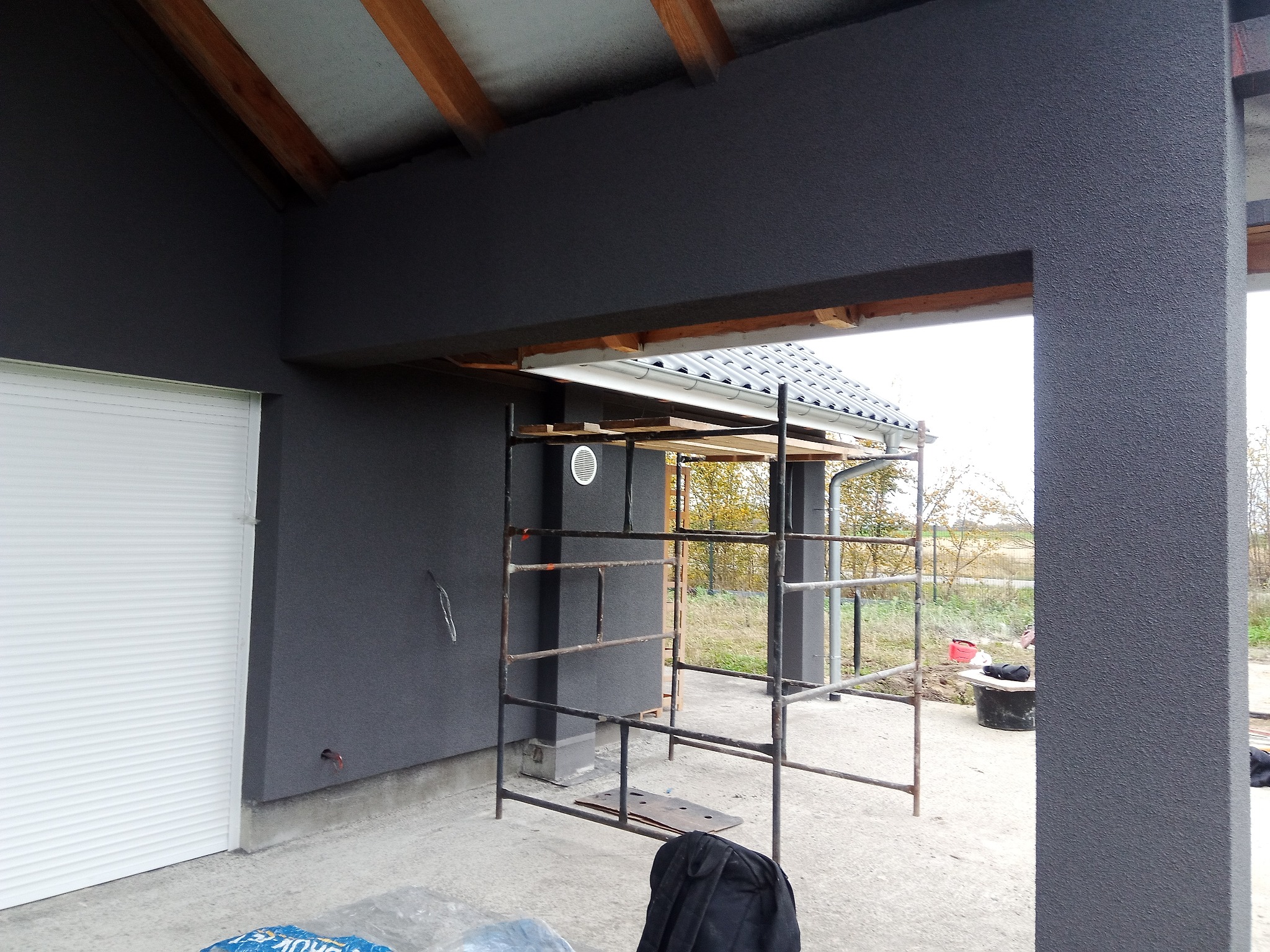 Szara elewacja domu w trakcie tynkowania, widoczne rusztowanie, boczna brama garażowa z roletą, drewniane elementy konstrukcyjne dachu.