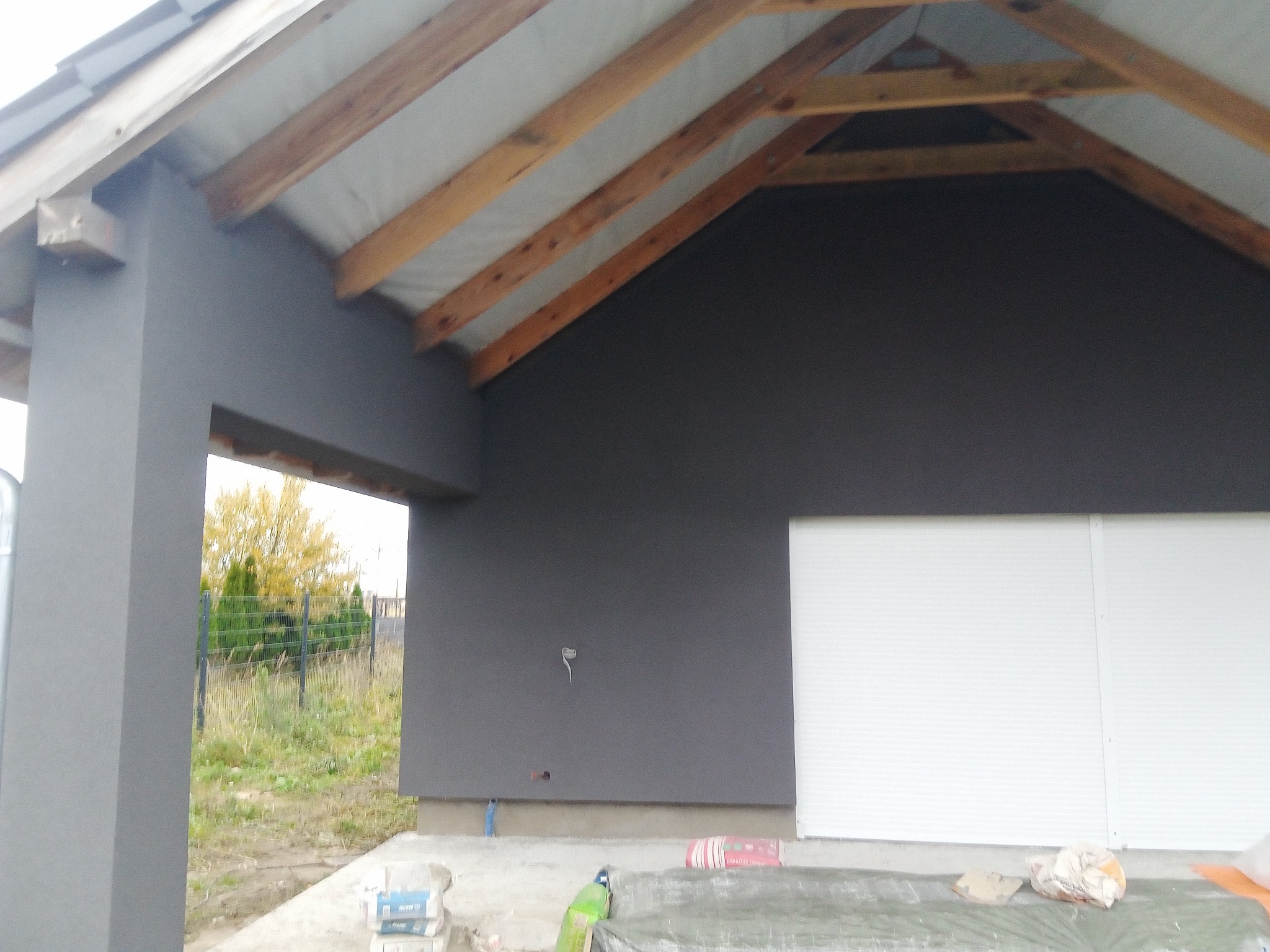 Szara elewacja budynku w trakcie tynkowania, widoczne drewniane elementy konstrukcyjne dachu, worki z zaprawą tynkarską na podłożu.