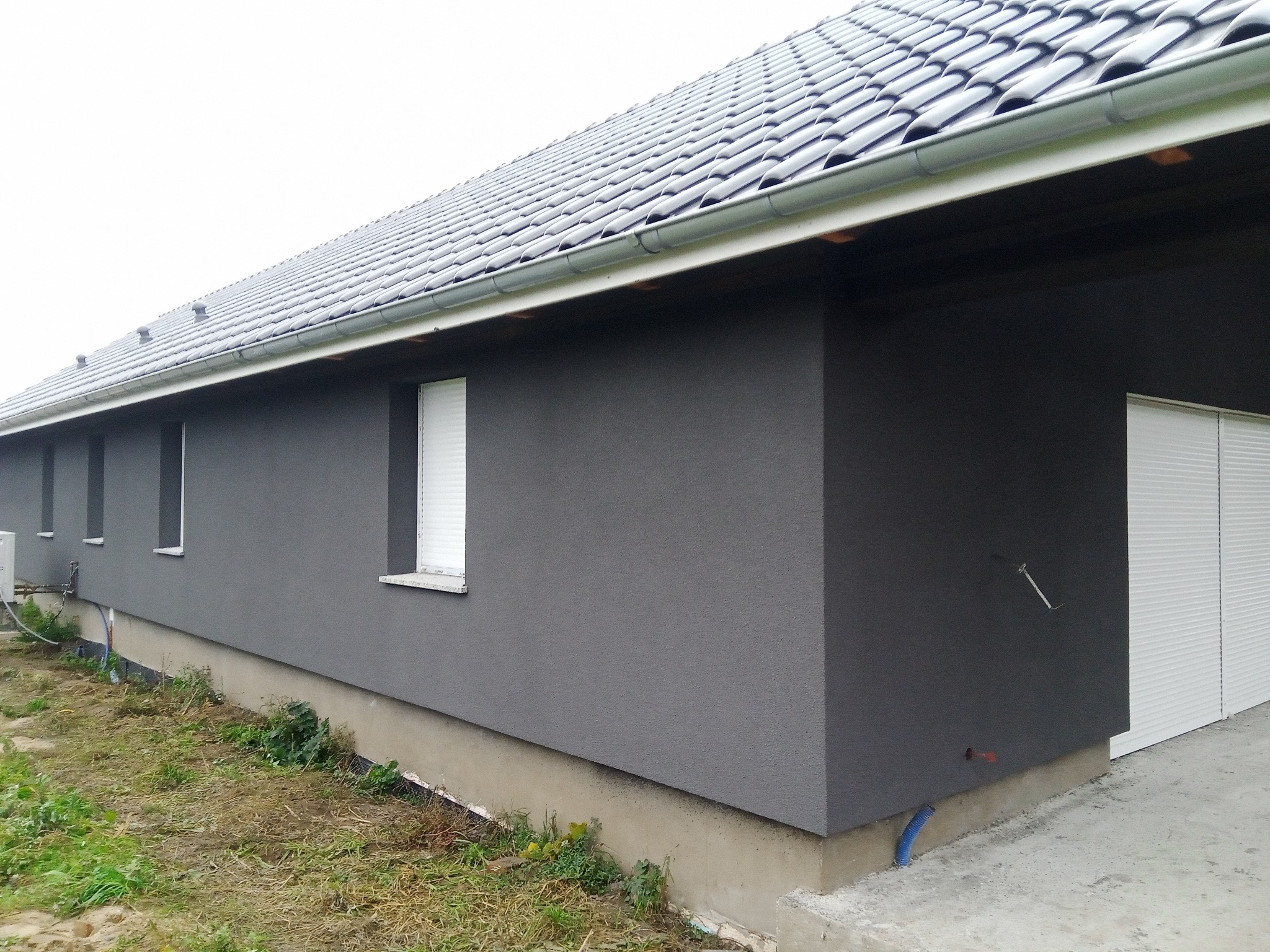 Szary dom jednorodzinny z ciemną elewacją i szarą dachówką, widoczne okna z białymi roletami i fragment niezagospodarowanego terenu.