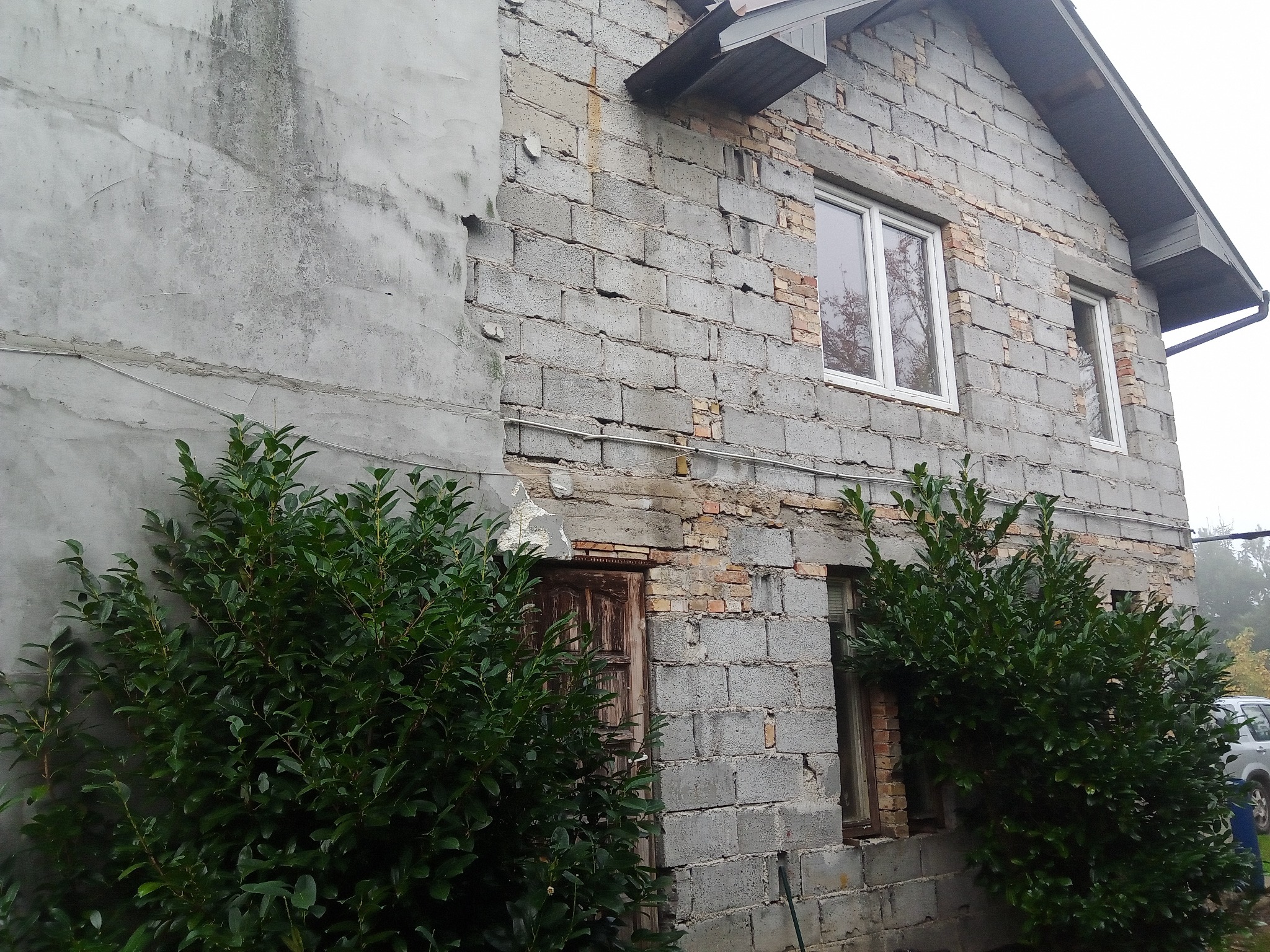 Niedokończona elewacja budynku z szarego betonu komórkowego i fragmentami starego tynku, widoczne uszkodzenia i brakujące elementy, okna w białych ramach, zarośnięte krzewami.