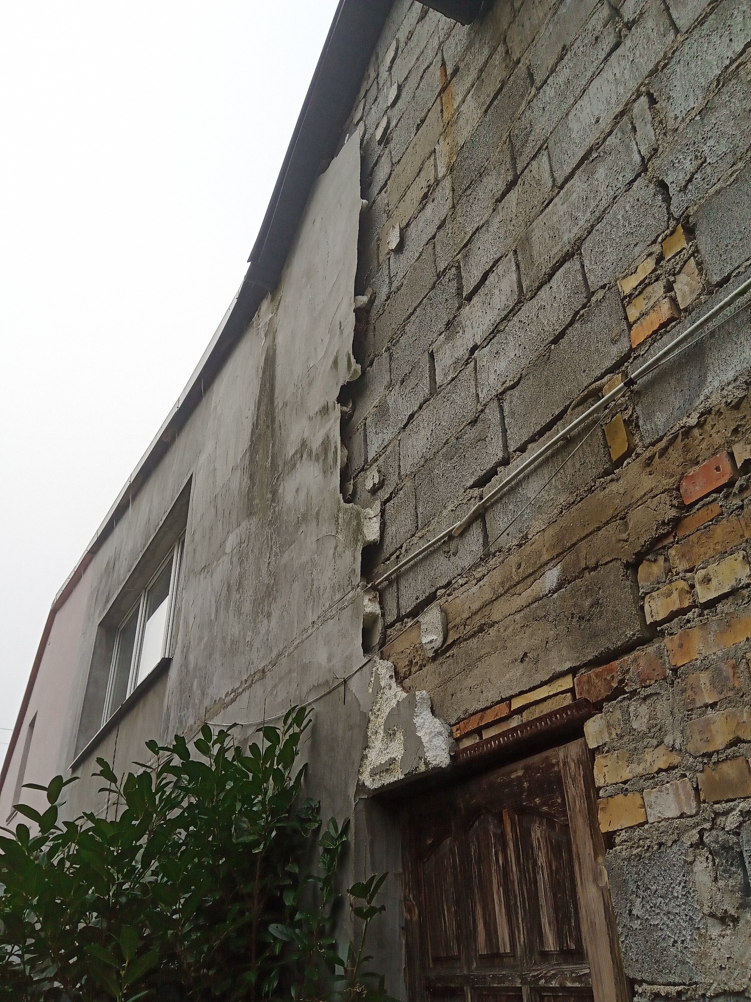 Narożnik budynku z odpadającym tynkiem i uszkodzoną elewacją z cegły, widoczne warstwy konstrukcyjne oraz zniszczone wejście.