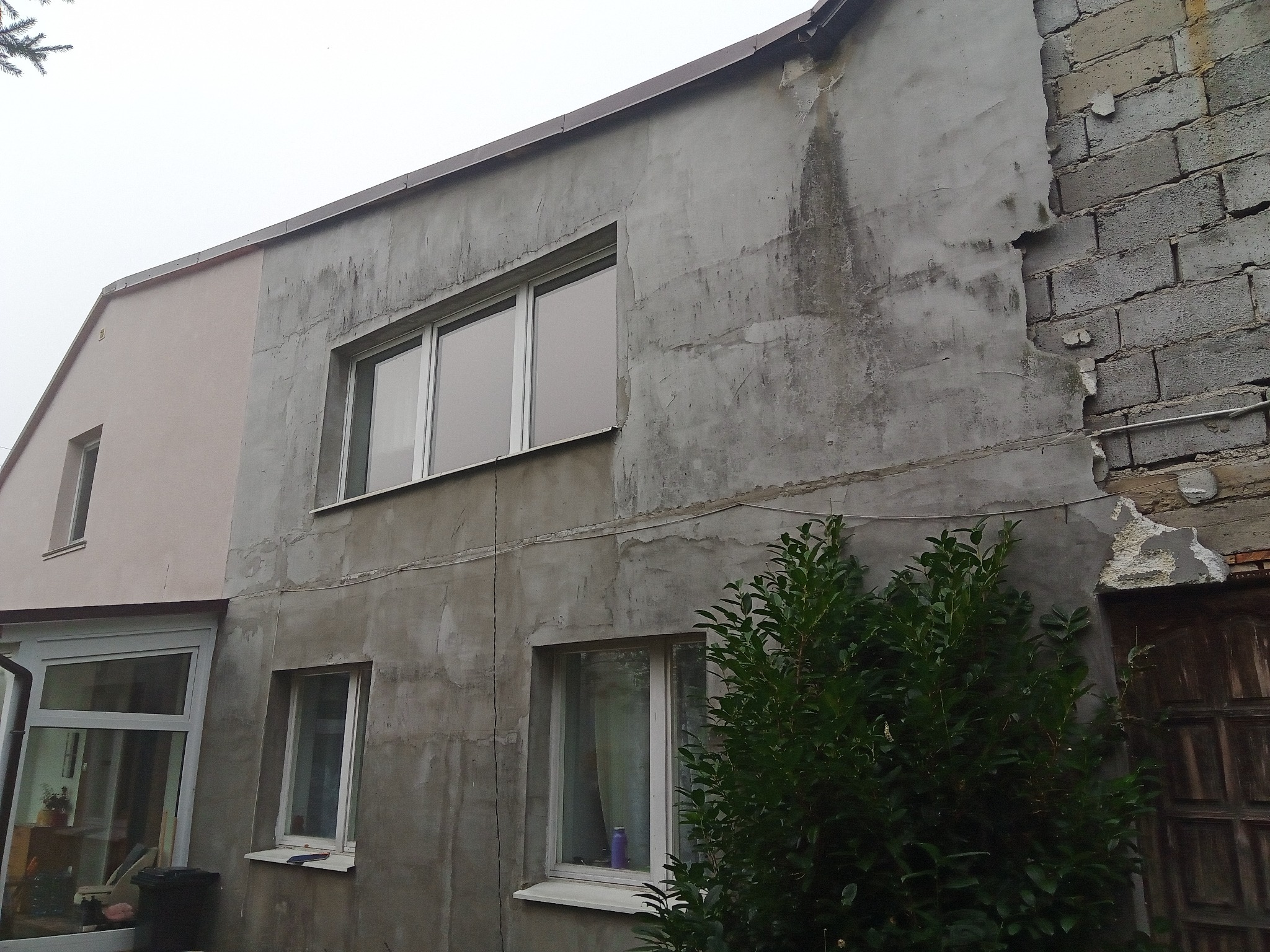 Niedokończona elewacja budynku z surowym tynkiem, widoczne fragmenty starej cegły i nowe okna PCV, krzew przed budynkiem.