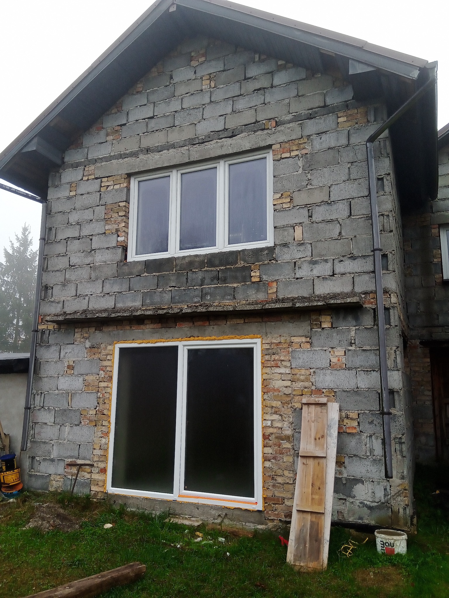 Surowa elewacja dwukondygnacyjnego domu z szarego bloczka, z widocznymi fragmentami cegieł, nowymi oknami i żółtą pianką montażową wokół dolnego okna, przy ścianie oparta deska, widoczne rynny.