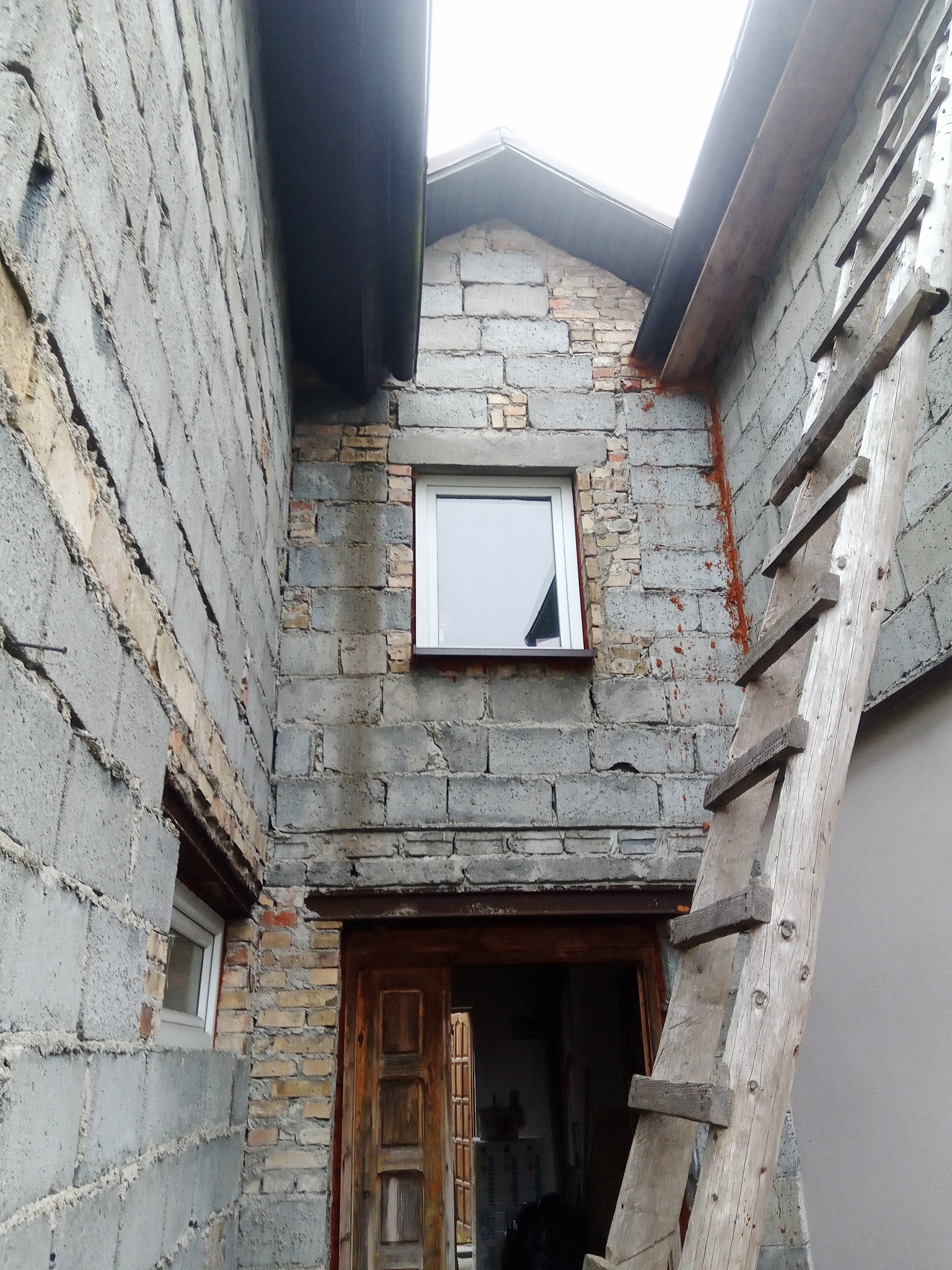 Surowa elewacja budynku z szarego bloczka betonowego i cegły, z drewnianą drabiną opartą o ścianę i widocznym wejściem do wnętrza.