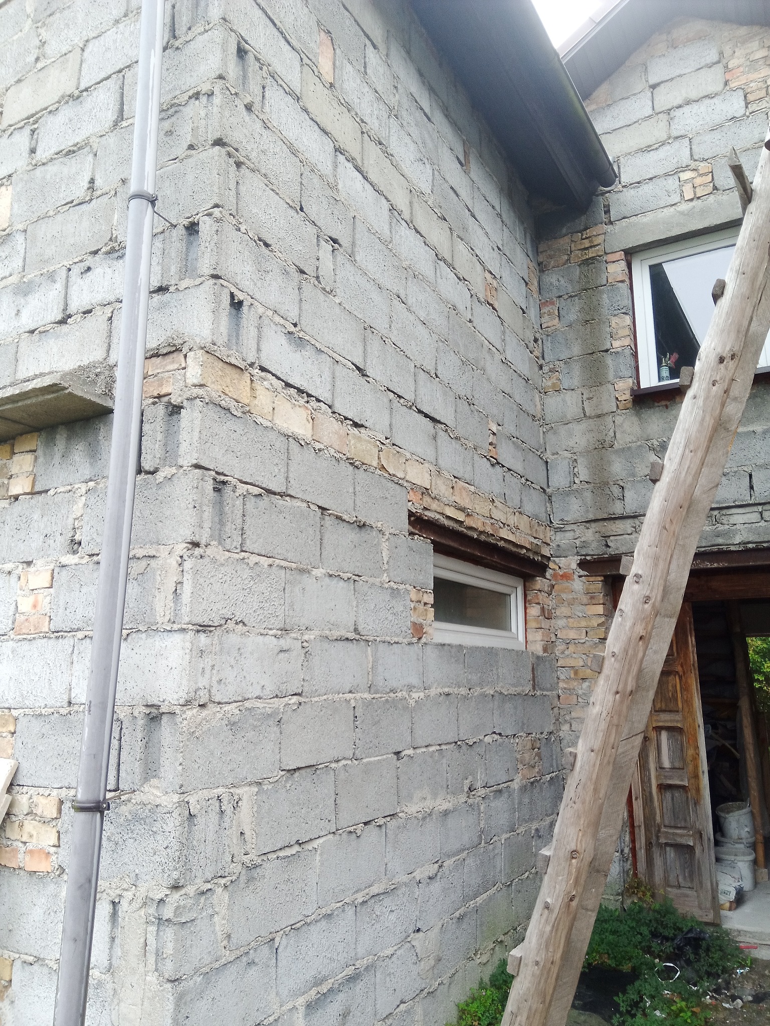 Surowa ściana budynku z szarego bloczka, fragmentami odsłonięta cegła, widoczne okna, rynna i drewniana drabina oparta o ścianę.