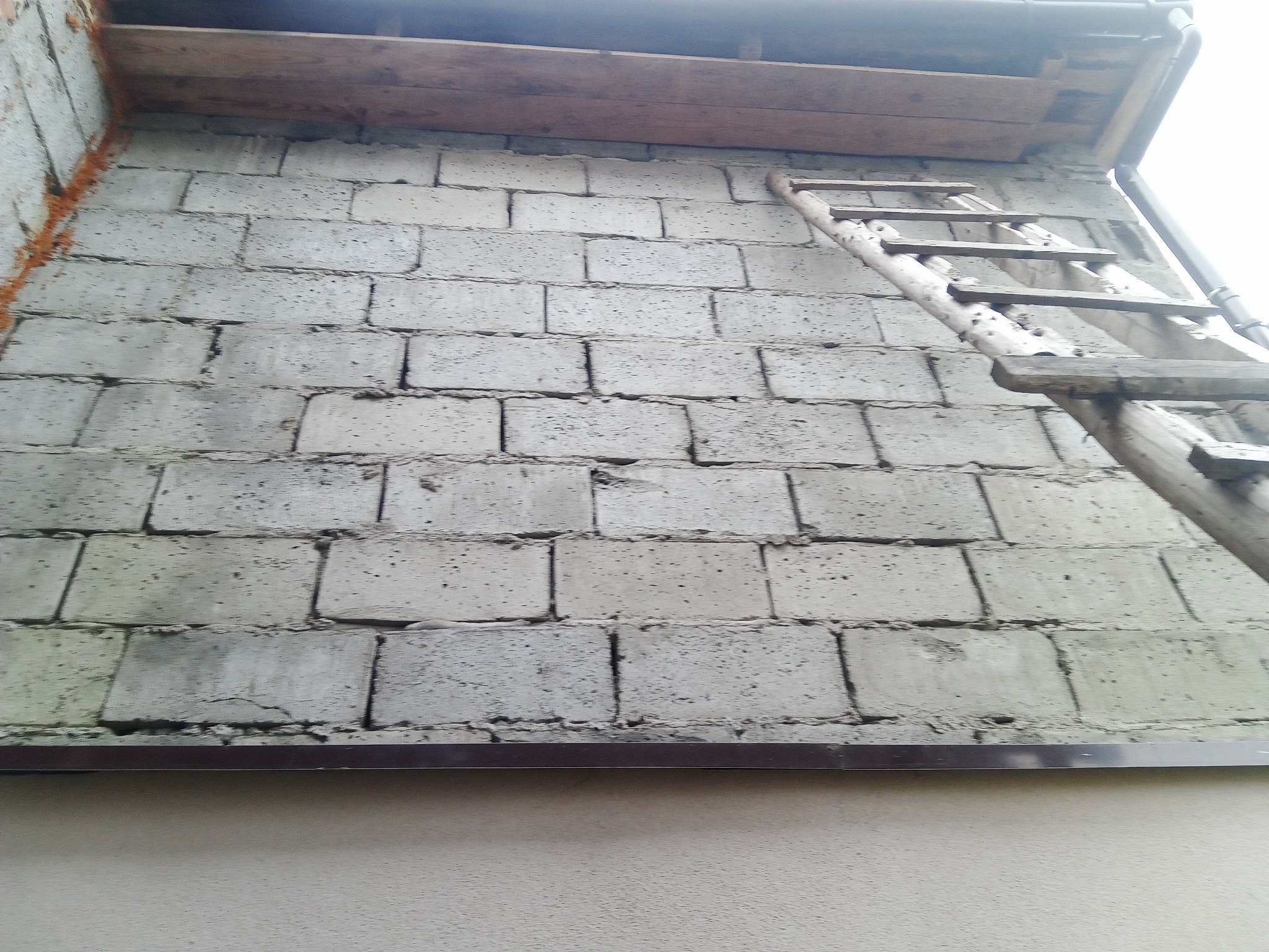 Surowa ściana z betonu komórkowego przed ociepleniem, widoczna drewniana drabina oparta o ścianę, fragment dachu i rynna