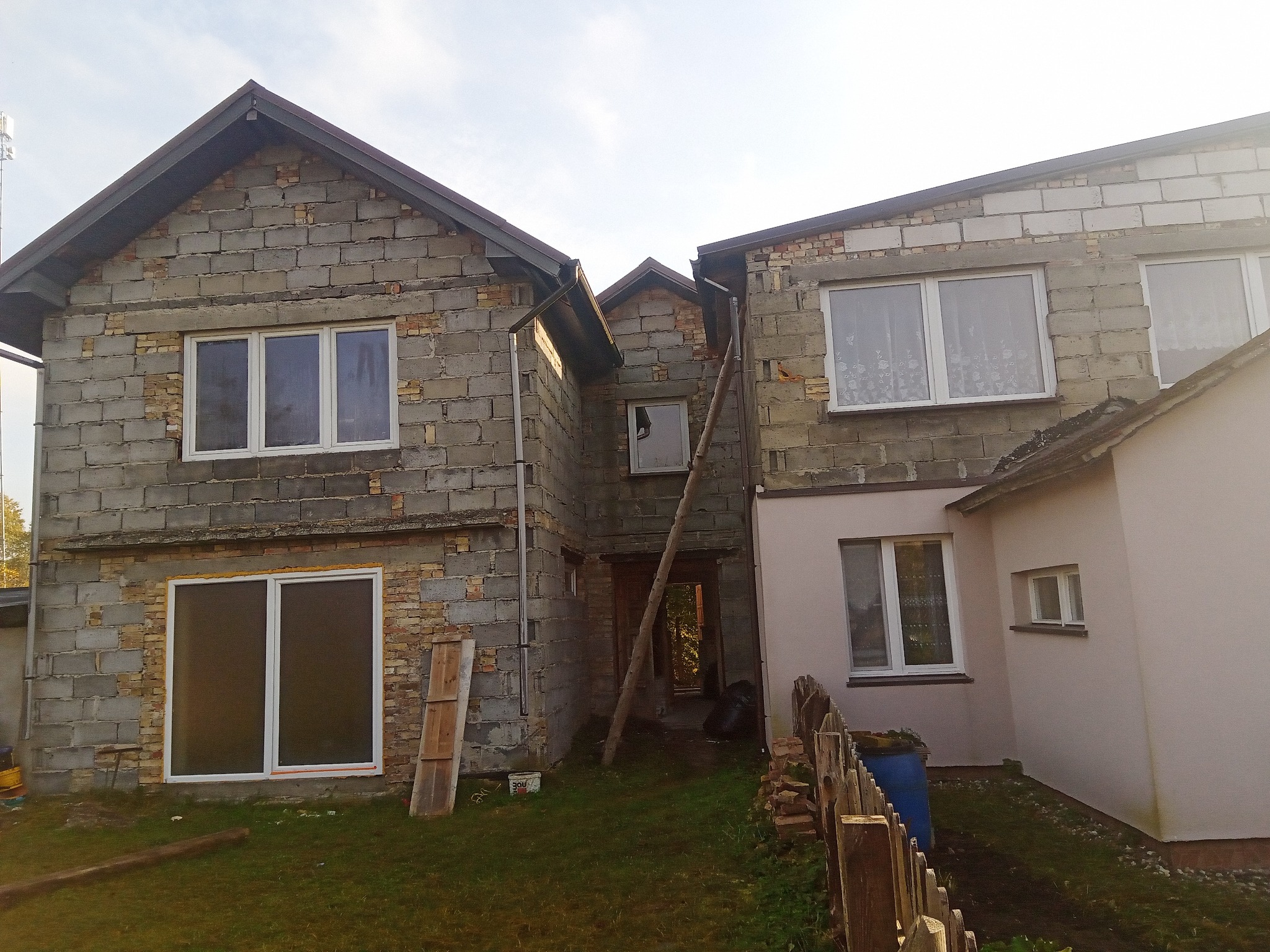 Surowa elewacja dwukondygnacyjnego domu w trakcie budowy, widoczne białe okna, szare bloczki i fragmenty cegieł, drewniane deski oparta o ścianę, teren wokół domu trawiasty.