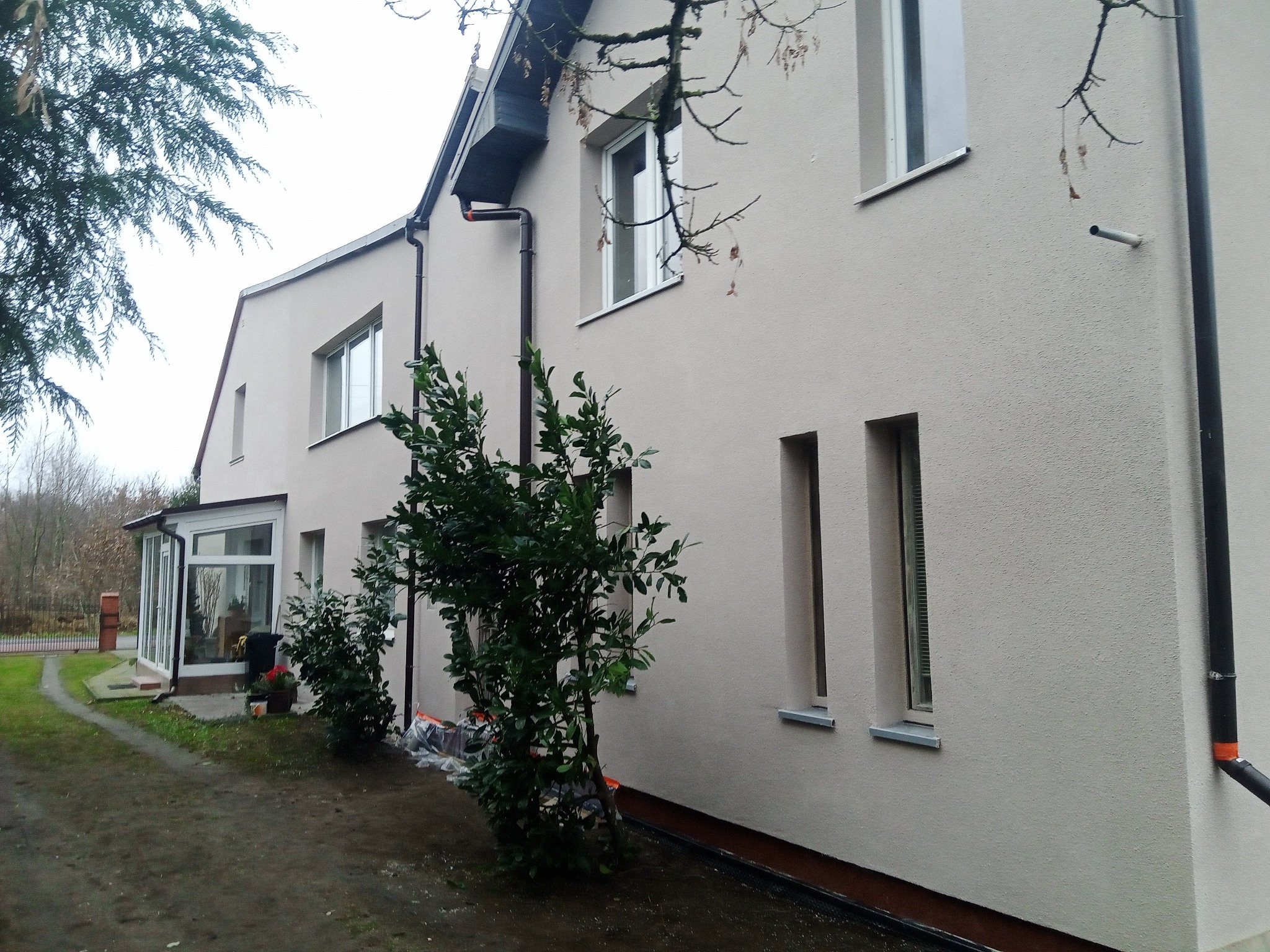 Jasna elewacja domu jednorodzinnego po tynkowaniu, widoczne okna z szarymi parapetami, czarne rynny i fragment ogrodu z zielenią