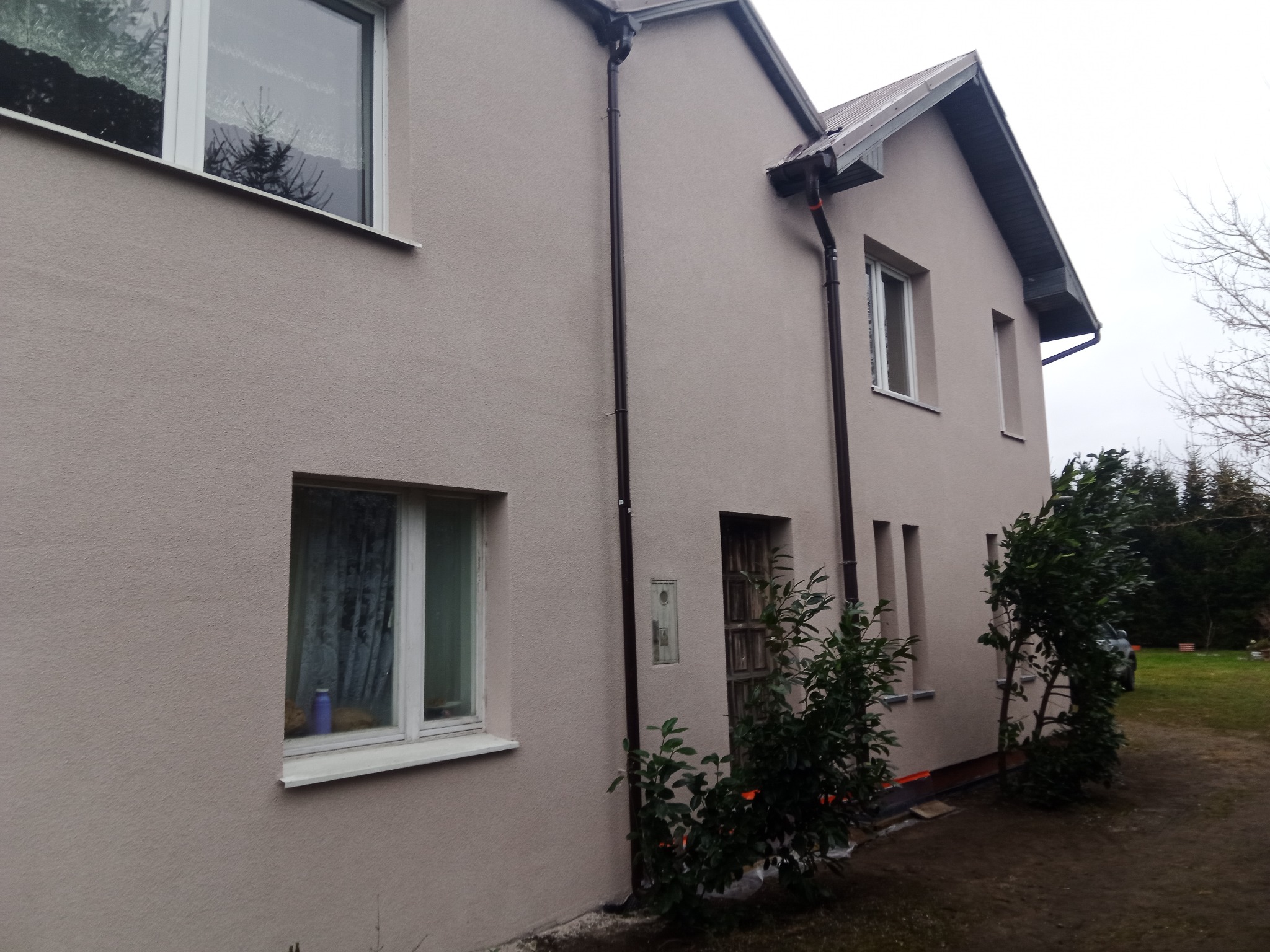 Jasna, otynkowana elewacja domu jednorodzinnego z brązowymi rynnami i białymi oknami; widoczne krzewy przy ścianie i fragment ogrodu