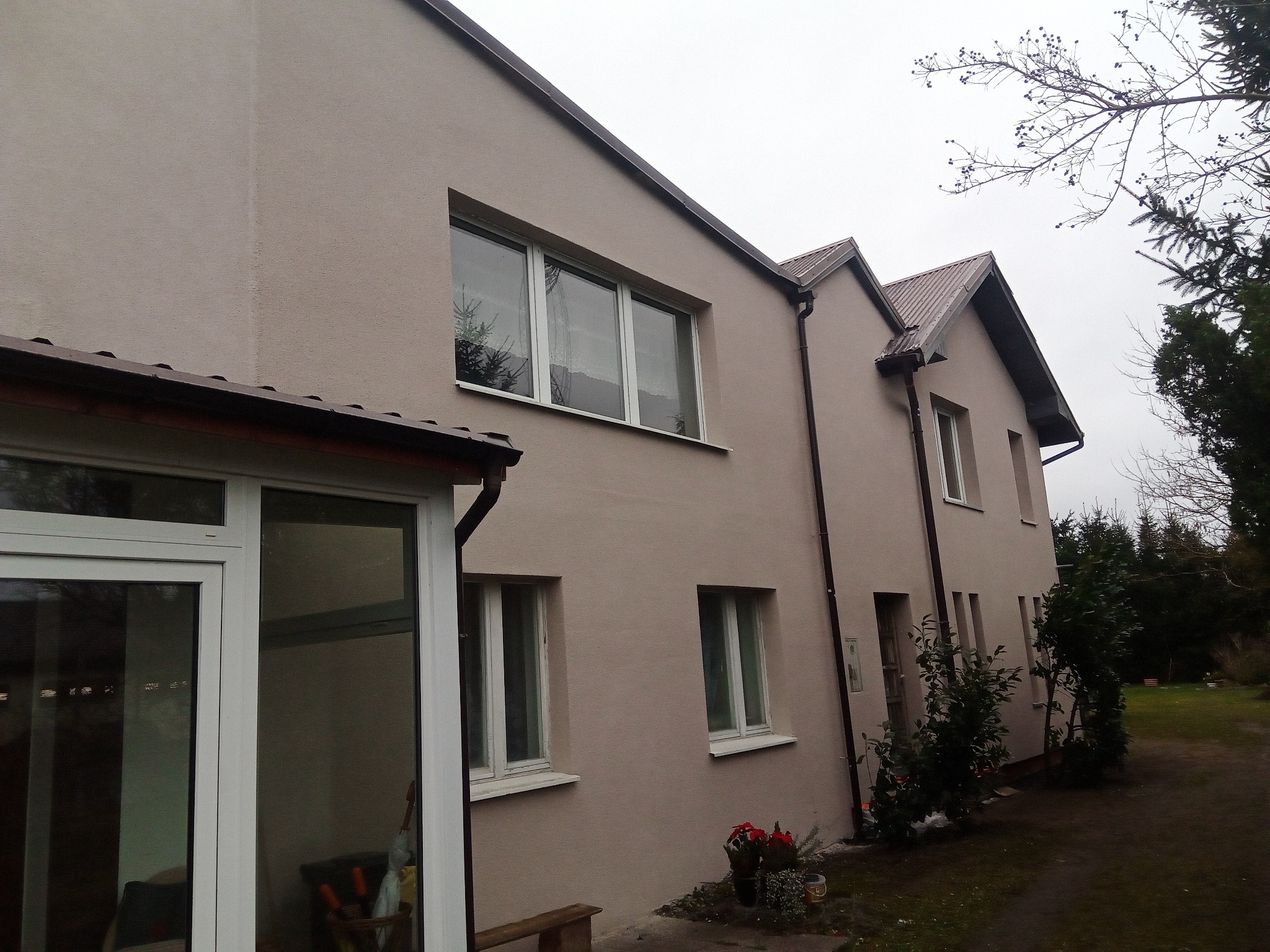 Dwukondygnacyjny dom jednorodzinny z beżową elewacją o strukturze baranka, widoczny z boku, z oknami PCV i brązowymi rynnami, otoczony zielenią.