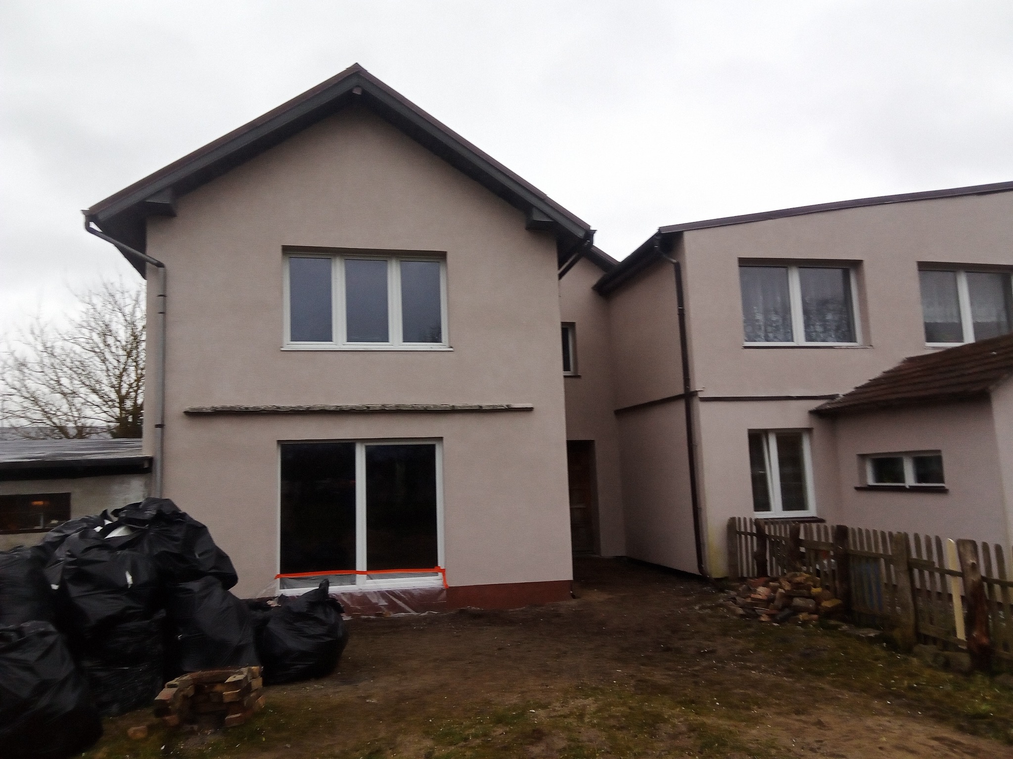 Dwa budynki po tynkowaniu elewacji w kolorze beżowym, z widocznymi workami z odpadami budowlanymi i folią ochronną przy oknie, sugerujące trwające prace wykończeniowe.