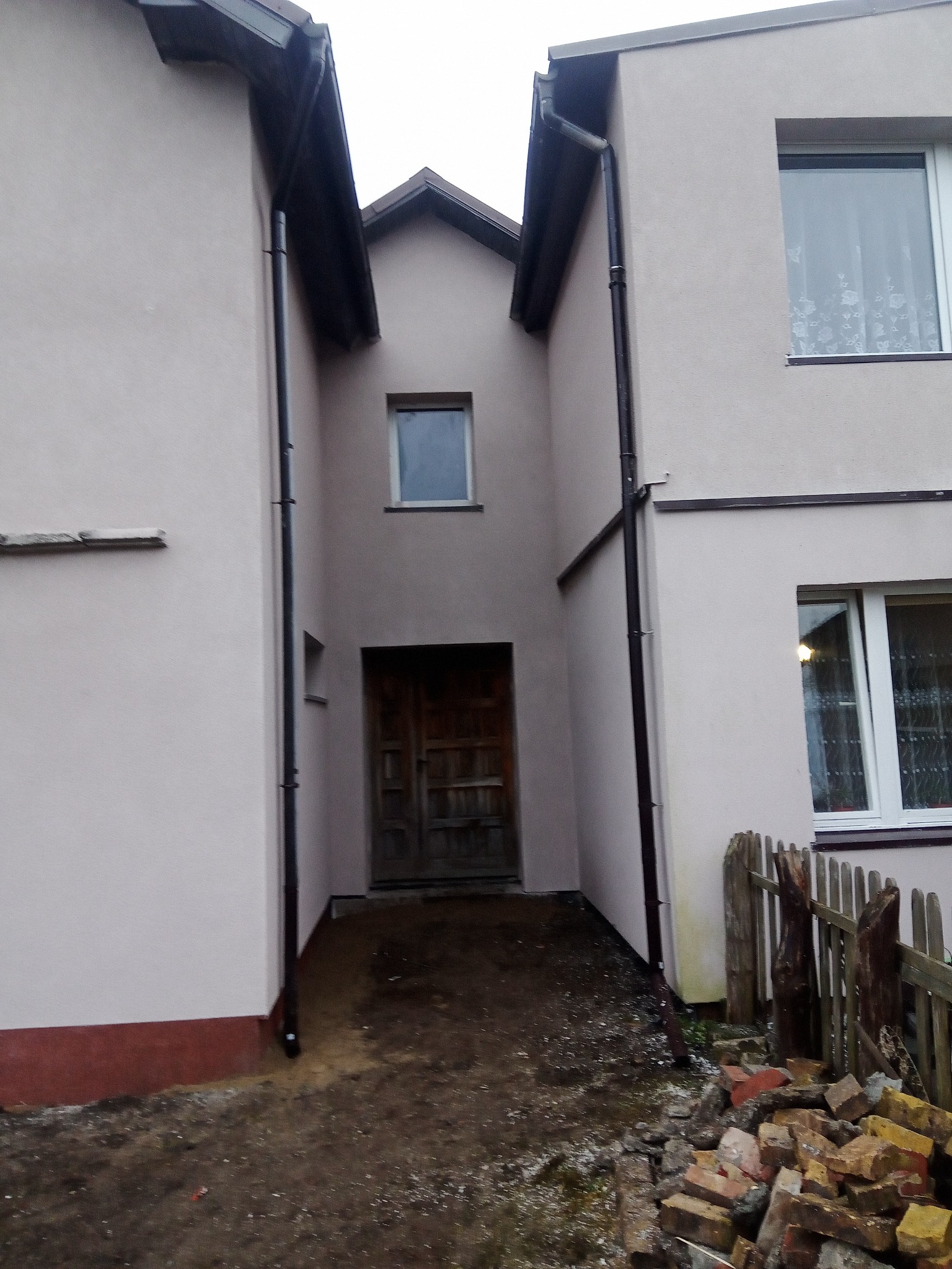 Elewacja budynku po renowacji z nowym tynkiem w odcieniach beżu, widoczne rynny, okna z firankami i fragment drewnianej bramy wejściowej, na pierwszym planie gruz ceglany i fragment płotu.