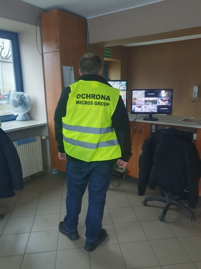 Pracownik ochrony w żółtej kamizelce z napisem 'OCHRONA MICROS GROUP' stoi tyłem do kamery przed monitorem z podglądem z kamer CCTV.