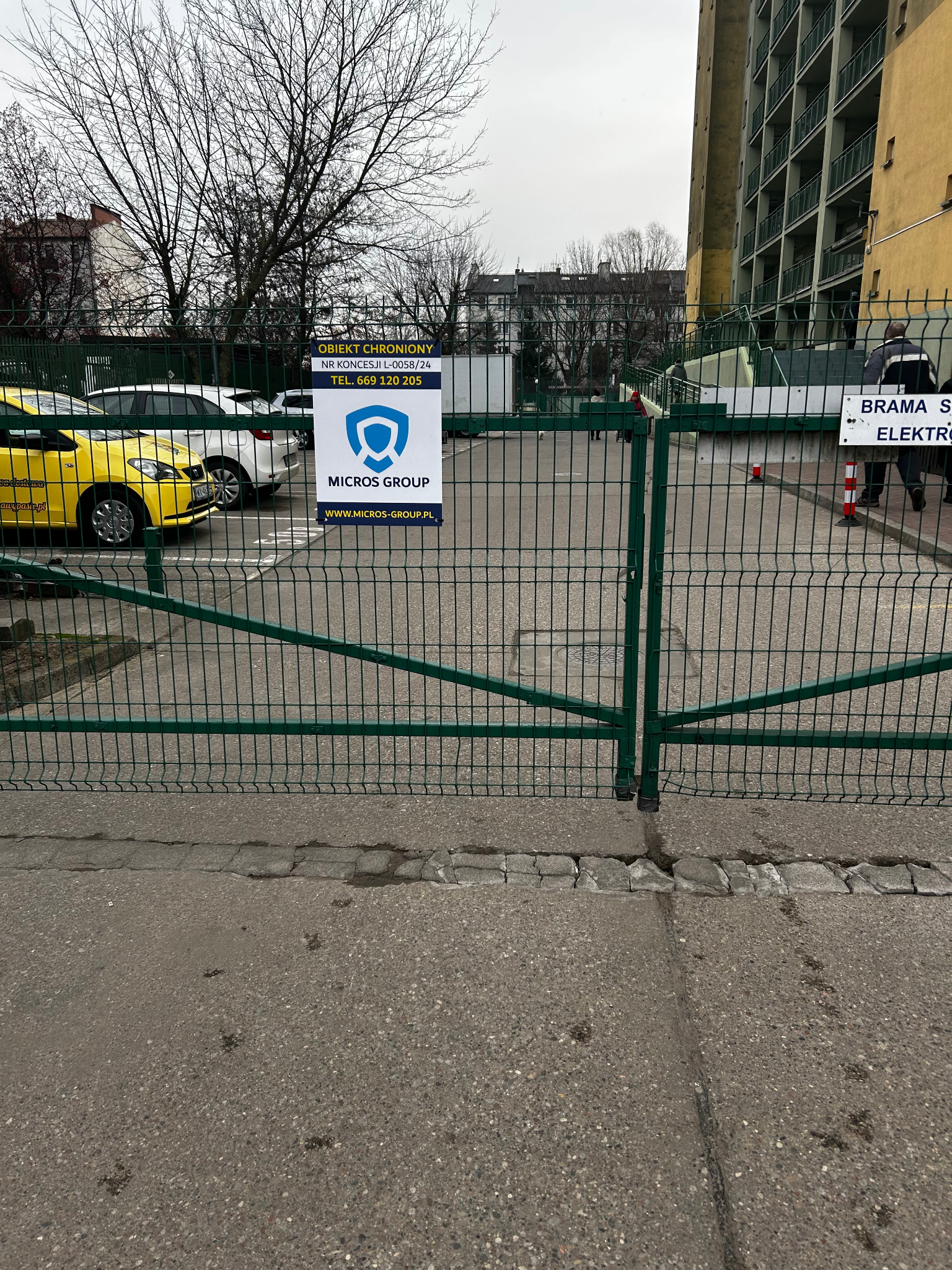 Zielona brama wjazdowa z tabliczką informującą o ochronie obiektu przez Micros Group, widoczny żółty samochód i budynek w tle.