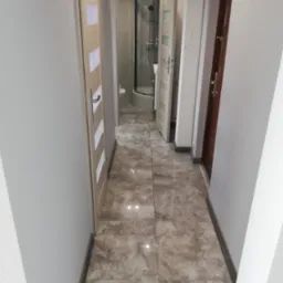 Korytarz z szarymi ścianami, płytkami imitującymi marmur na podłodze, widok na łazienkę z kabiną prysznicową i dwoma parami drzwi po bokach, jedne z drewnianymi wstawkami.