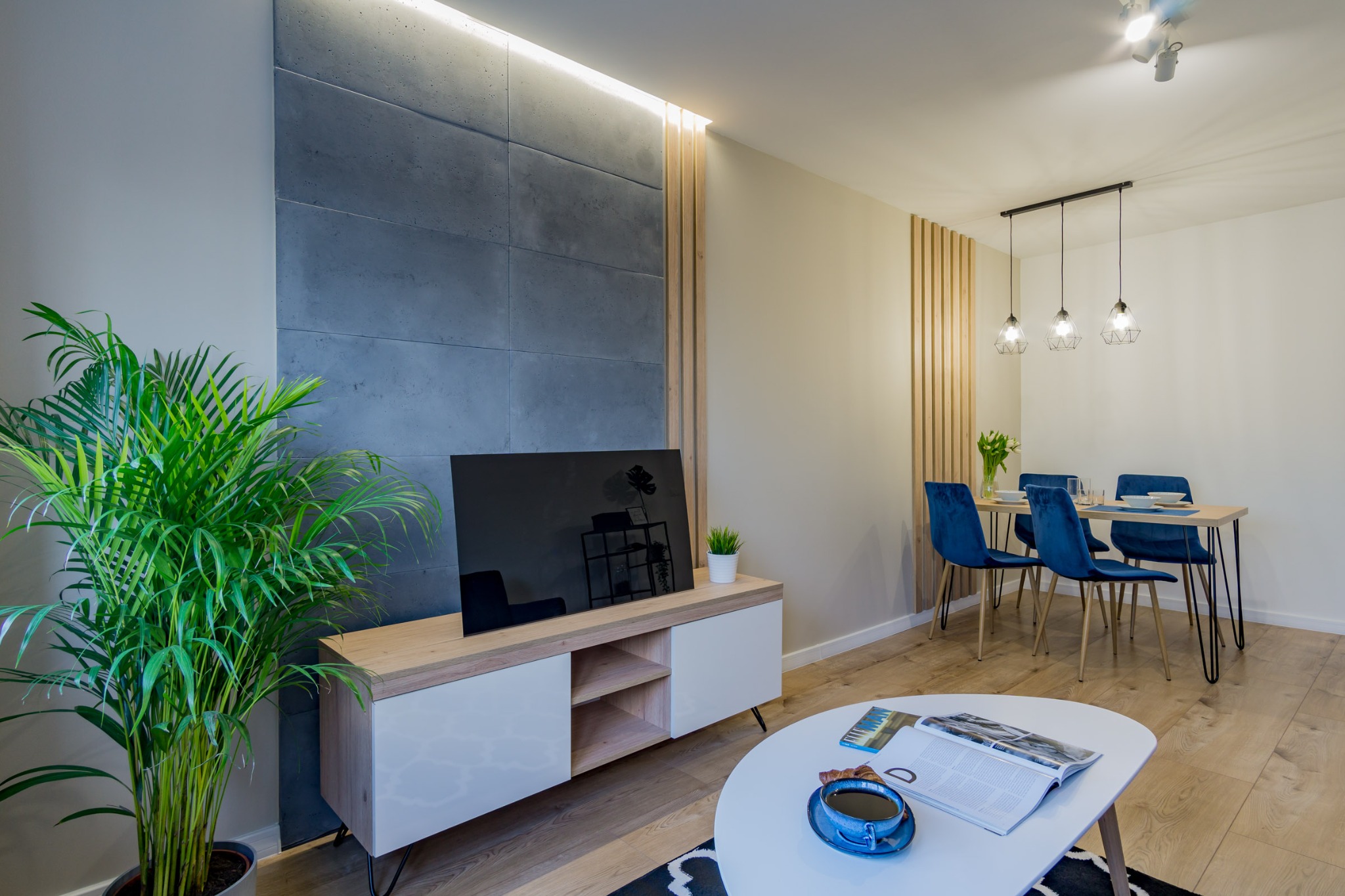 Nowoczesne wnętrze mieszkania z betonową ścianą, drewnianymi lamelami, parkietem, rośliną doniczkową i niebieskimi krzesłami przy stole.