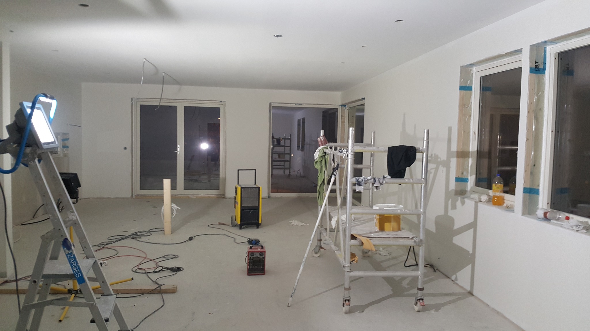 Wnętrze w trakcie malowania ścian na biało, widoczne okna zabezpieczone folią, drabina, rusztowanie, osuszacz powietrza, kable i lampa budowlana.