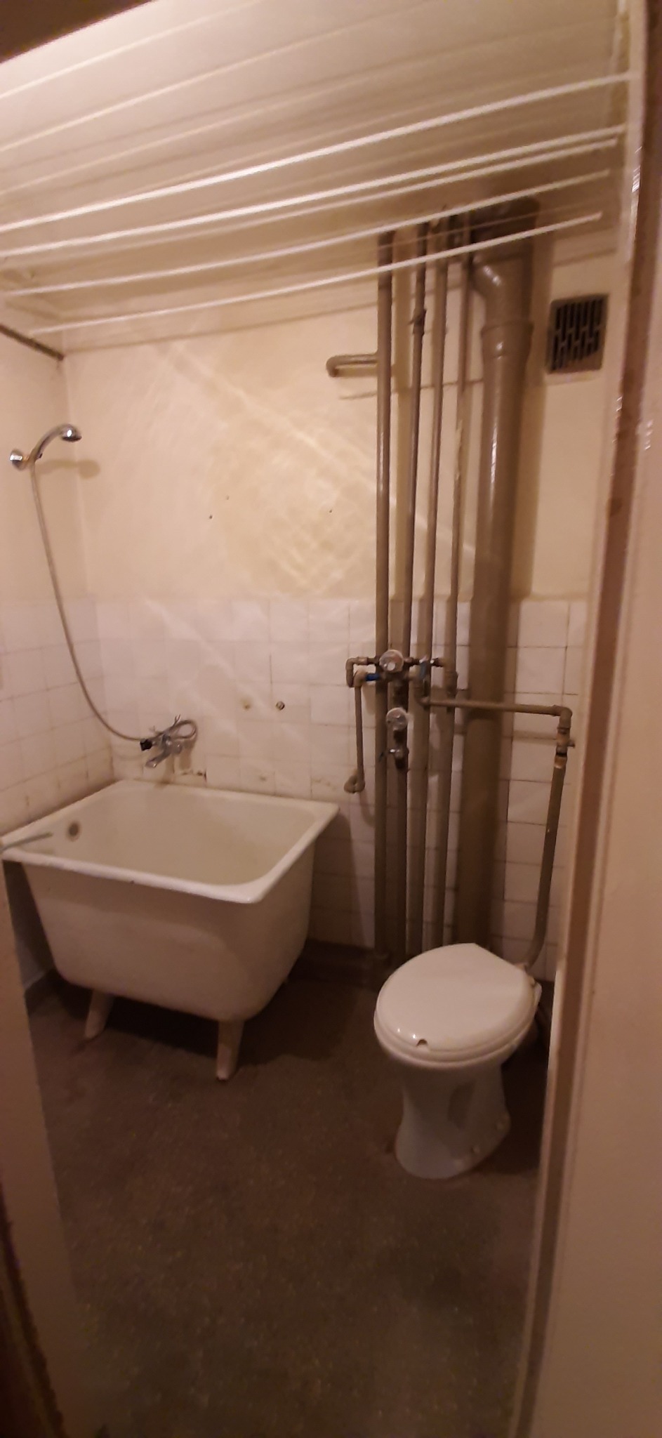 Niefotogeniczna łazienka z białą wanną na nóżkach, toaletą i widocznymi rurami instalacyjnymi.