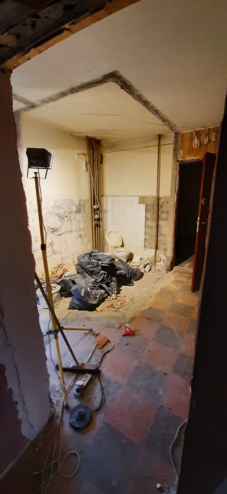 Łazienka w trakcie demontażu starej glazury i przygotowania do remontu, widoczne worki z gruzem, lampa budowlana na trójnogu oraz odłączona toaleta.
