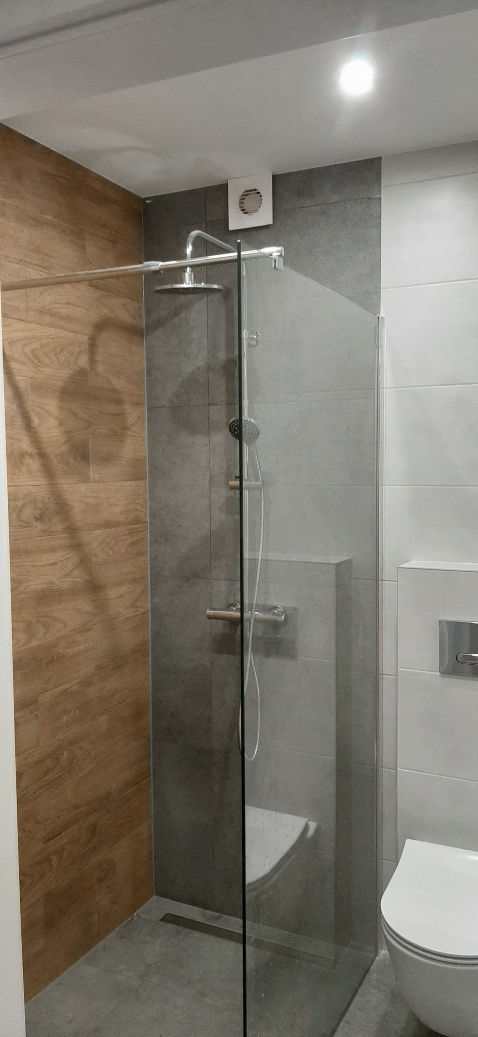 Nowoczesna kabina prysznicowa ze szklaną ścianką, deszczownicą i odpływem liniowym, obok wisząca toaleta i wentylator w szarej płytce.