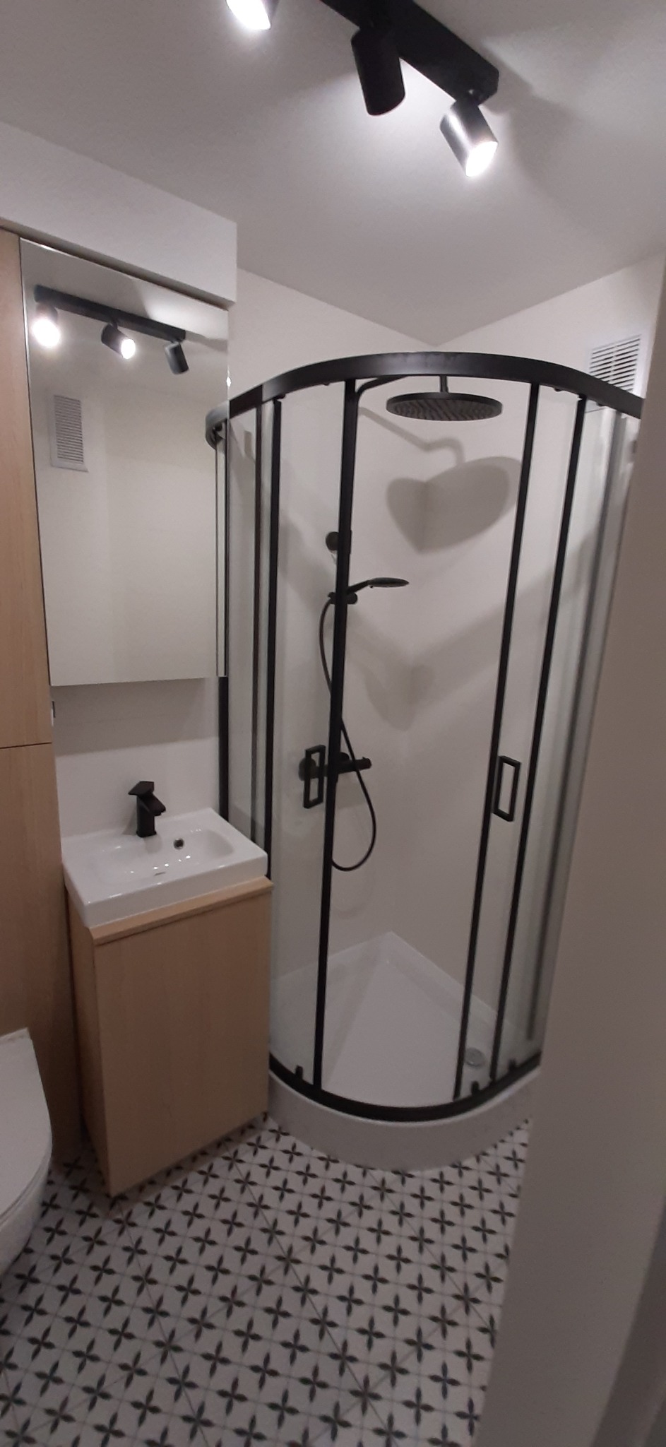 Nowoczesna łazienka z kabiną prysznicową z czarnymi profilami, umywalką z szafką w kolorze jasnego drewna i wzorzystą podłogą.