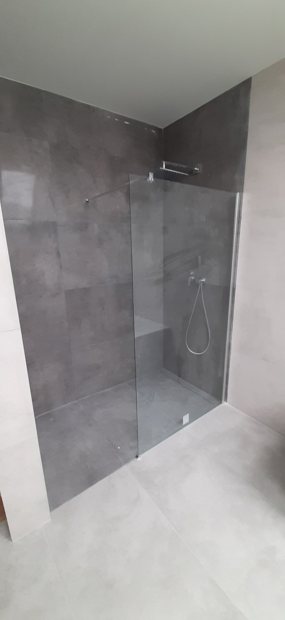 Nowoczesna kabina prysznicowa z deszczownicą i półką wewnątrz, wykończona płytkami w odcieniach szarości, z widoczną armaturą i szklaną ścianką.