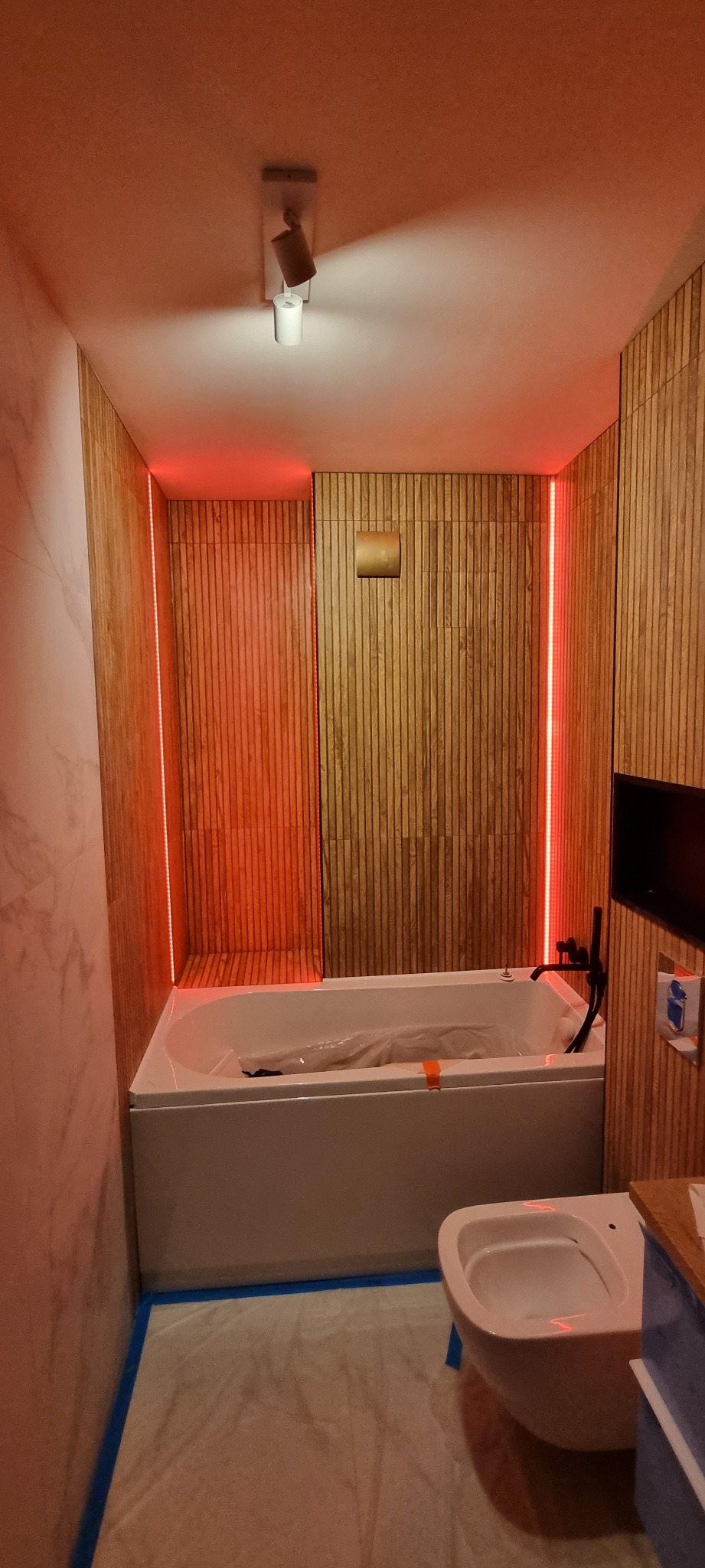 Nowoczesna łazienka w trakcie wykańczania, z drewnianą okładziną ścian, podświetleniem LED, białą wanną zabezpieczoną folią oraz wiszącą toaletą. Podłoga przykryta folią ochronną.