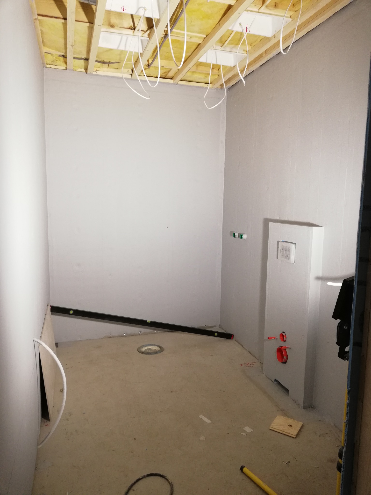 Łazienka w trakcie remontu: widoczne drewniane belki sufitu z izolacją, szare ściany po gruntowaniu, zabudowa stelaża podtynkowego WC z czerwonymi rurami, poziomica na ścianie, otwór odpływowy...