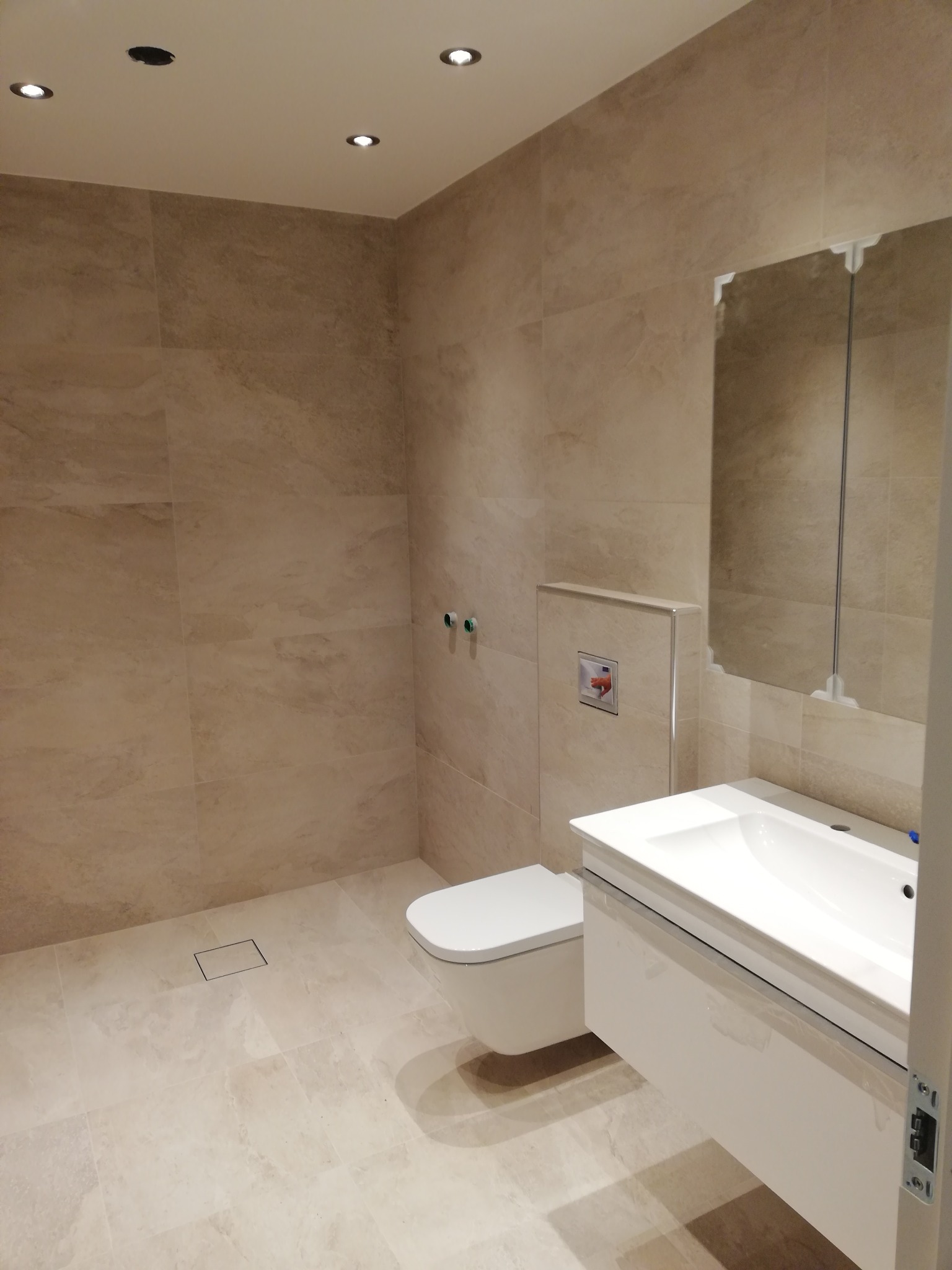 Jasna łazienka z beżowymi płytkami imitującymi marmur, z podwieszaną toaletą, szafką z umywalką i lustrem z szafką, oświetlenie punktowe w suficie.