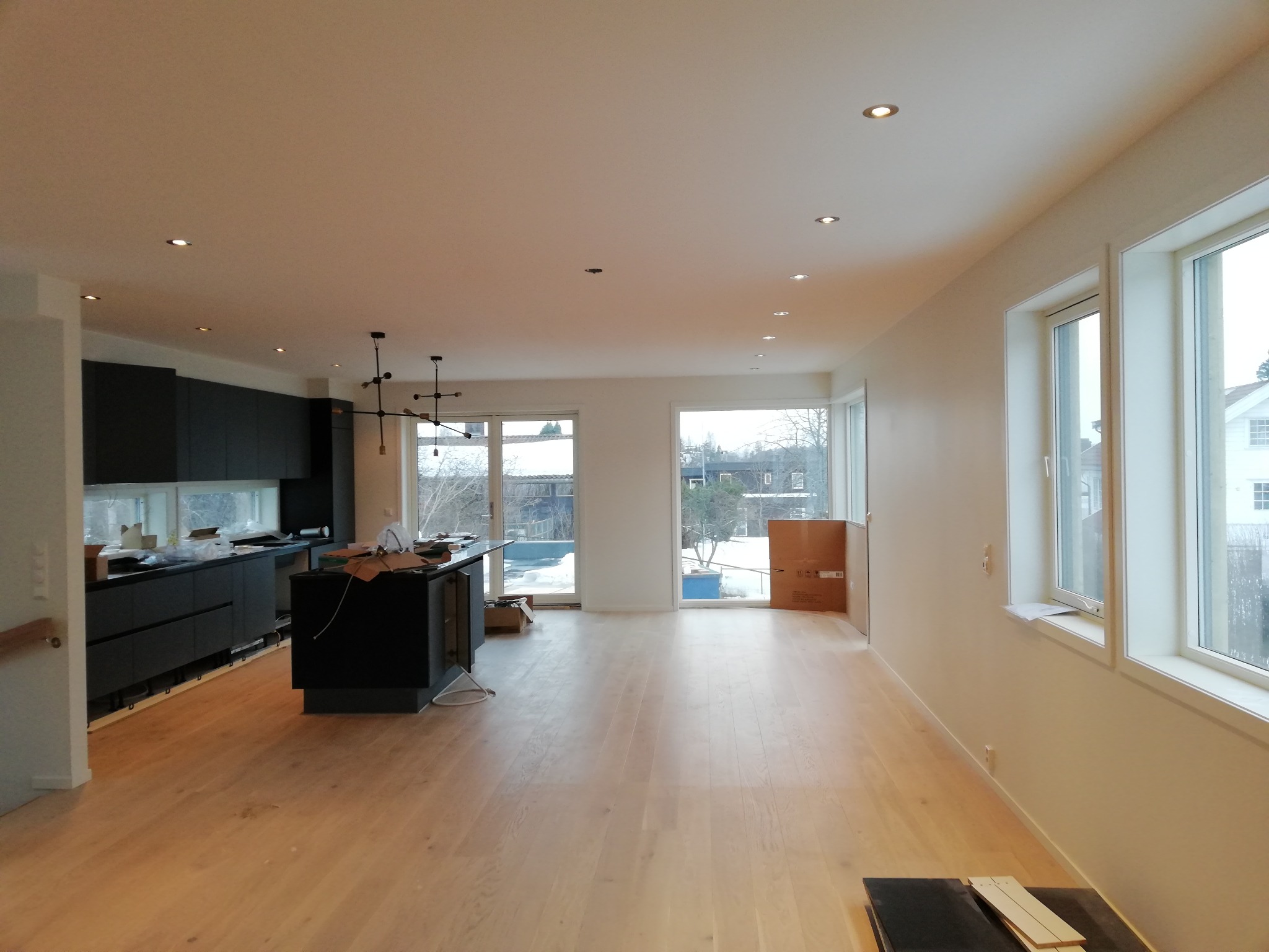 Przestronne wnętrze domu z jasną drewnianą podłogą, nowoczesną czarną kuchnią z wyspą, dużymi oknami i sufitowymi lampami punktowymi. Częściowo widoczne kartony i przedmioty sugerują trwające prace...