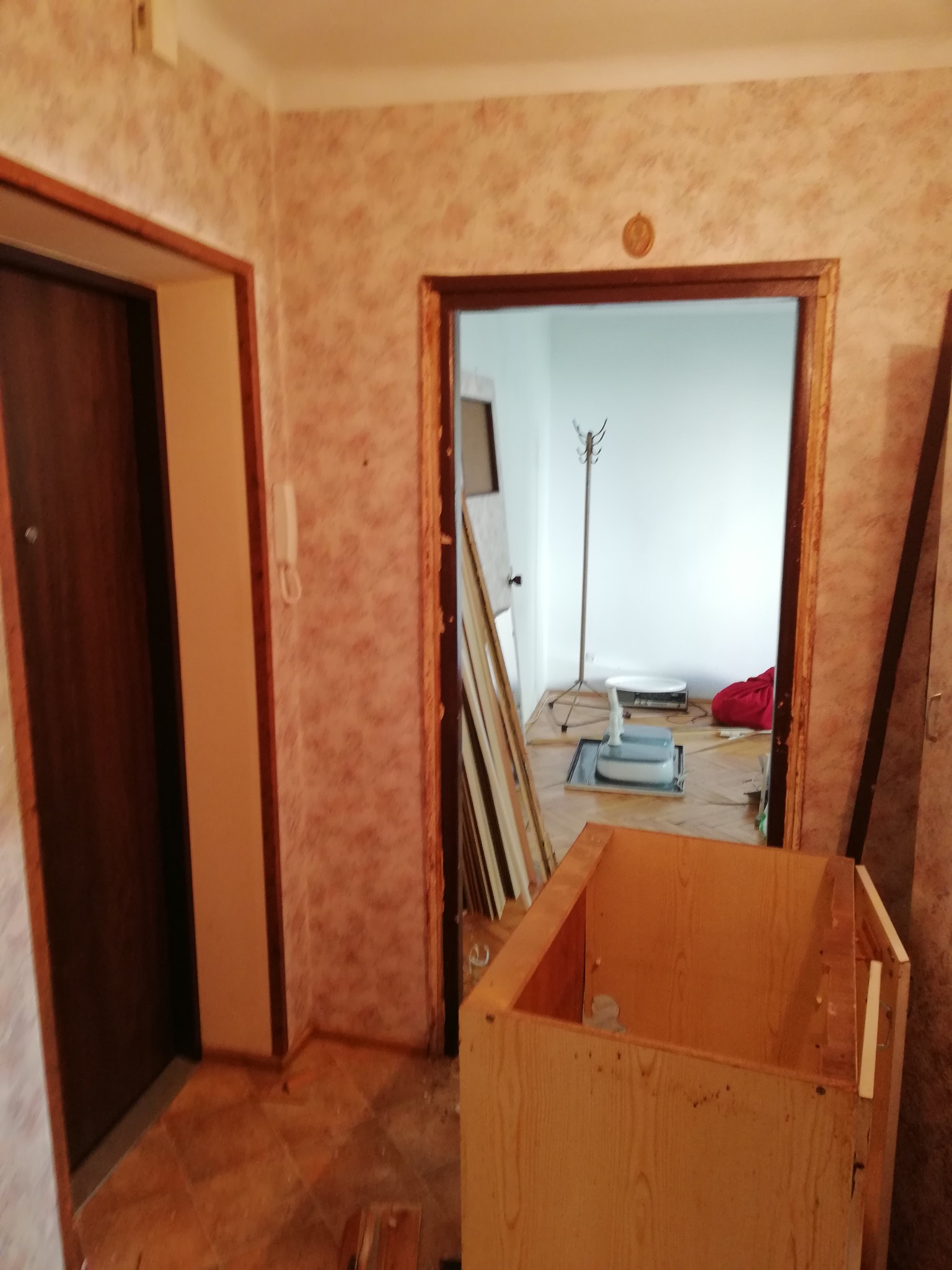 Wnętrze mieszkania w trakcie demontażu starej zabudowy, widoczny fragment demontowanej szafki z płyty wiórowej, odsłonięte futryny drzwiowe i fragment ściany z tapetą.